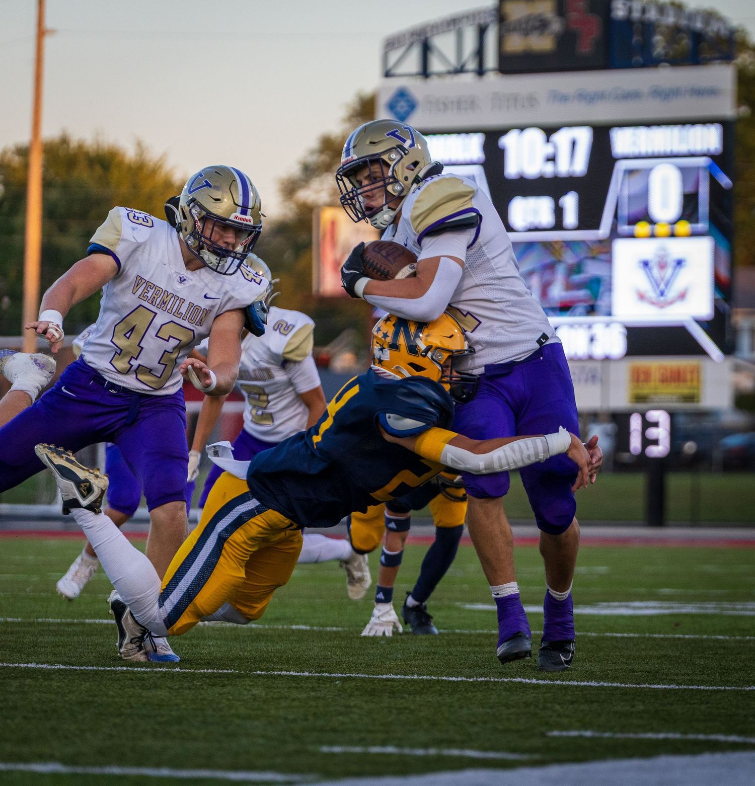 Purple and gold football players battle during a high school game with scoreboard showing 10:17 in background.