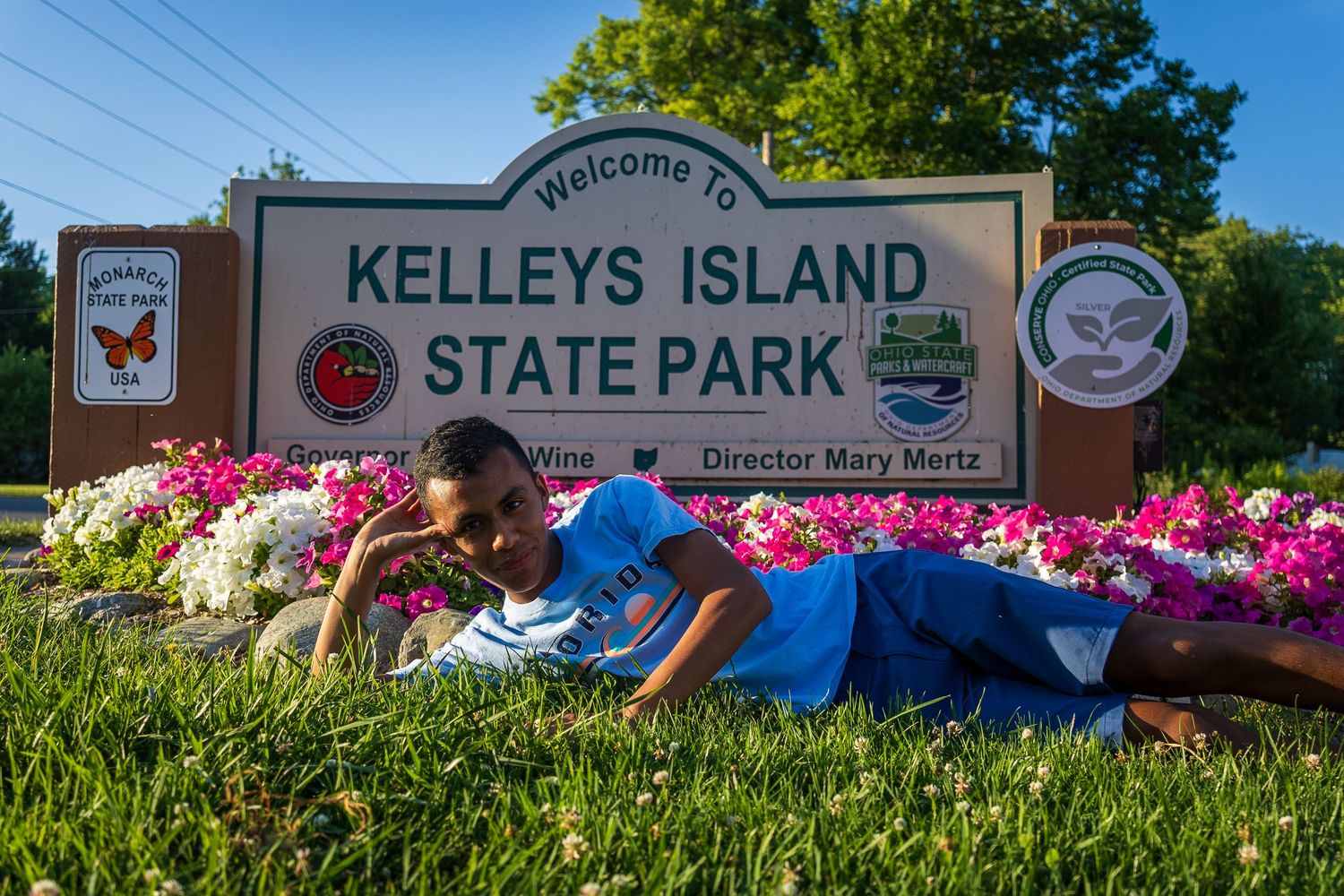Child poses in front of Kelleys Island State Park welcome sign surrounded by colorful flowers on a sunny day.