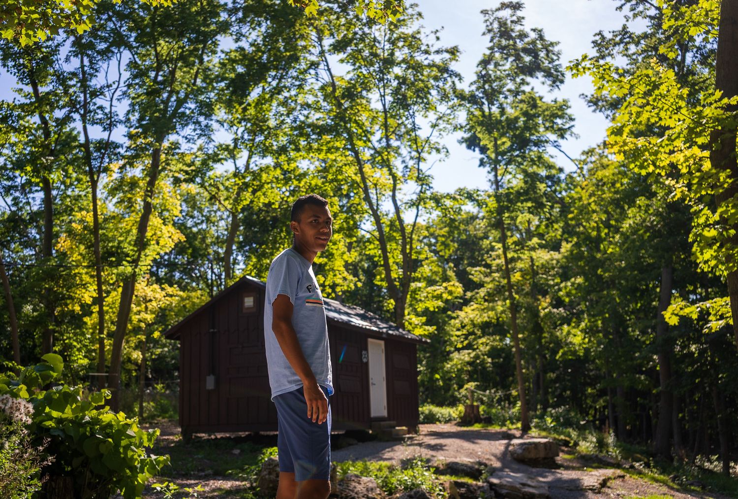 Man stands in wooded area near rustic brown cabin surrounded by lush green trees in summer.