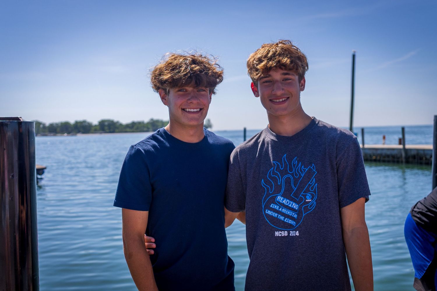 Two teenage boys with curly hair smiling together at sunny waterfront dock.