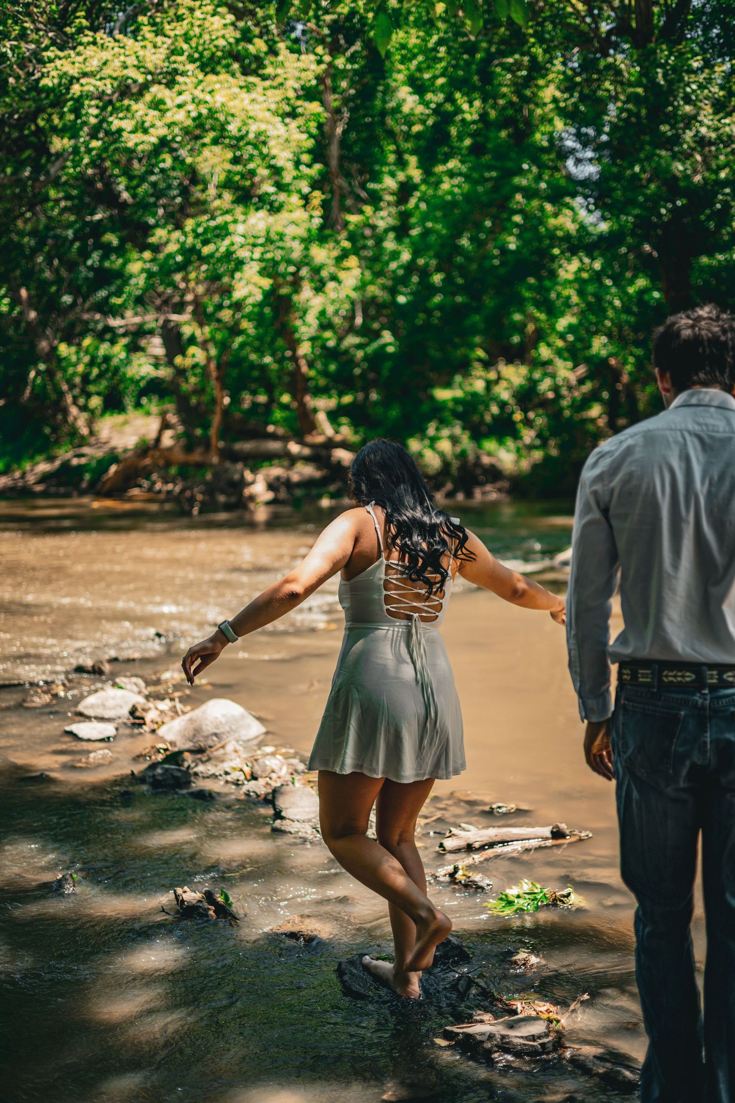 Couple walks hand-in-hand barefoot through shallow creek surrounded by lush green forest trees.
