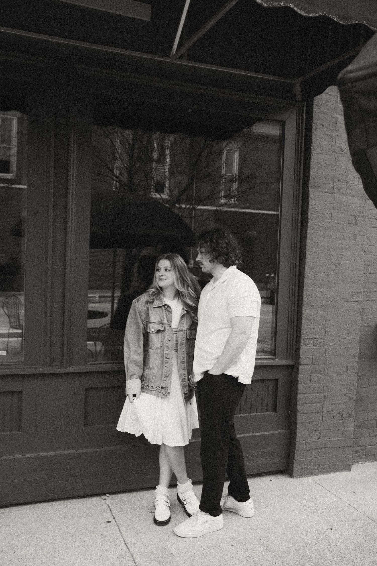 Black and white photo of couple holding hands and smiling outside a brick building storefront.