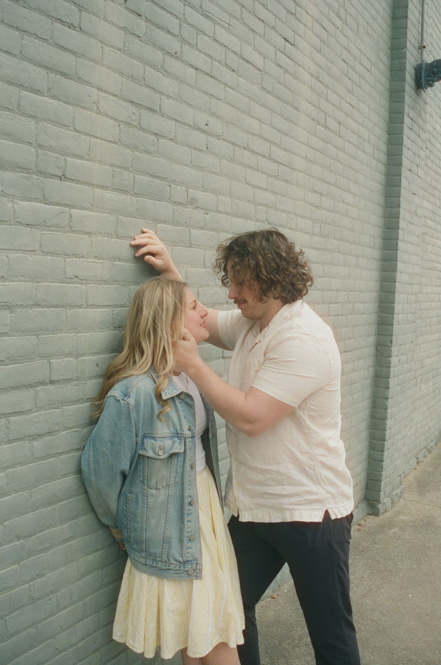 Couple in casual clothes sharing an intimate moment leaning against a painted gray brick wall.