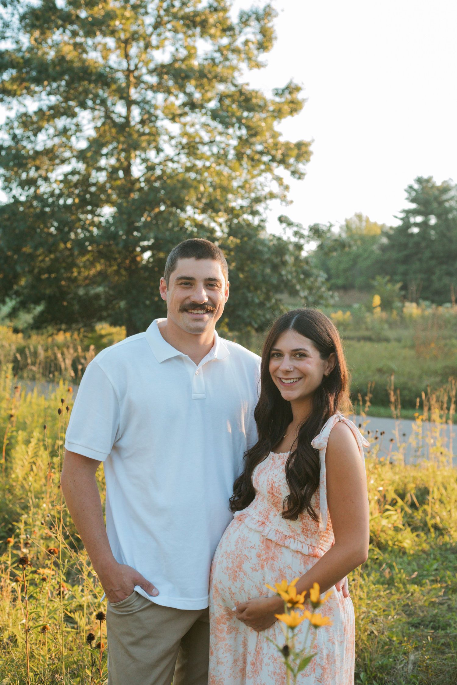 Smiling couple poses in a sunny wildflower field, woman in floral maternity dress cradling baby bump.