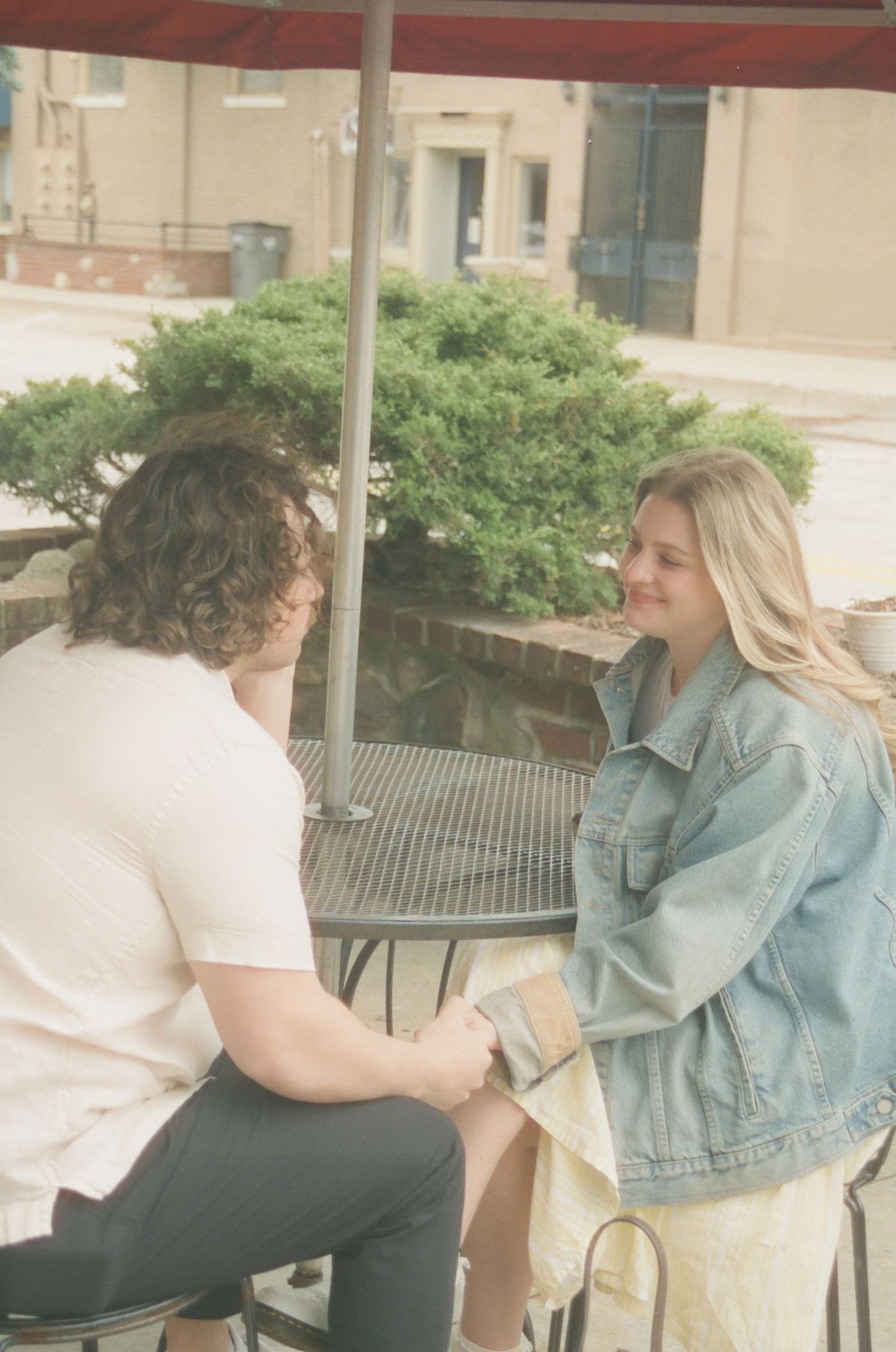 Couple holding hands at outdoor cafe table, woman in denim jacket and man in white shirt enjoying a date.