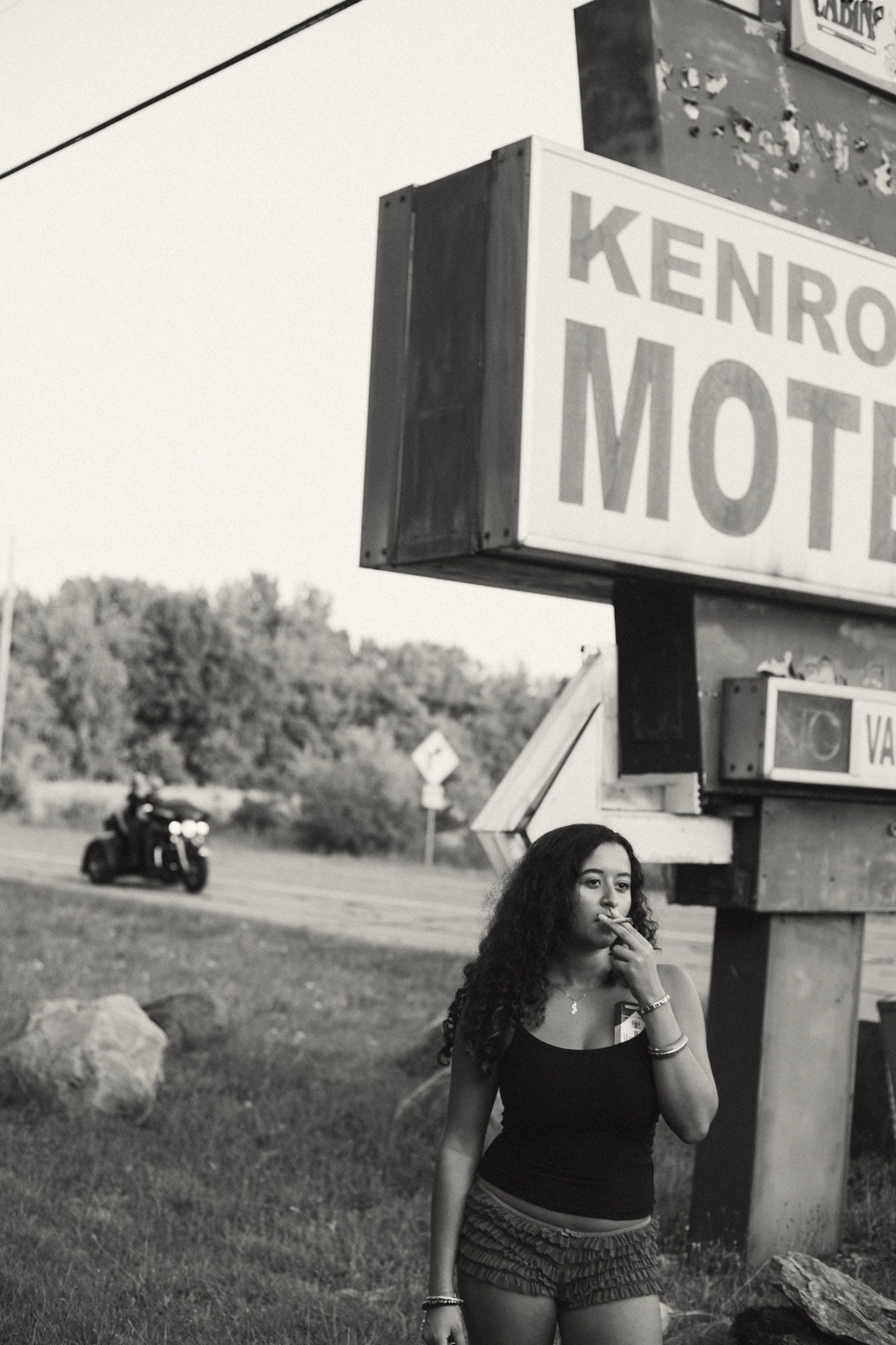 Black and white photo of woman with curly hair standing beneath Kenro Motel sign outdoors.