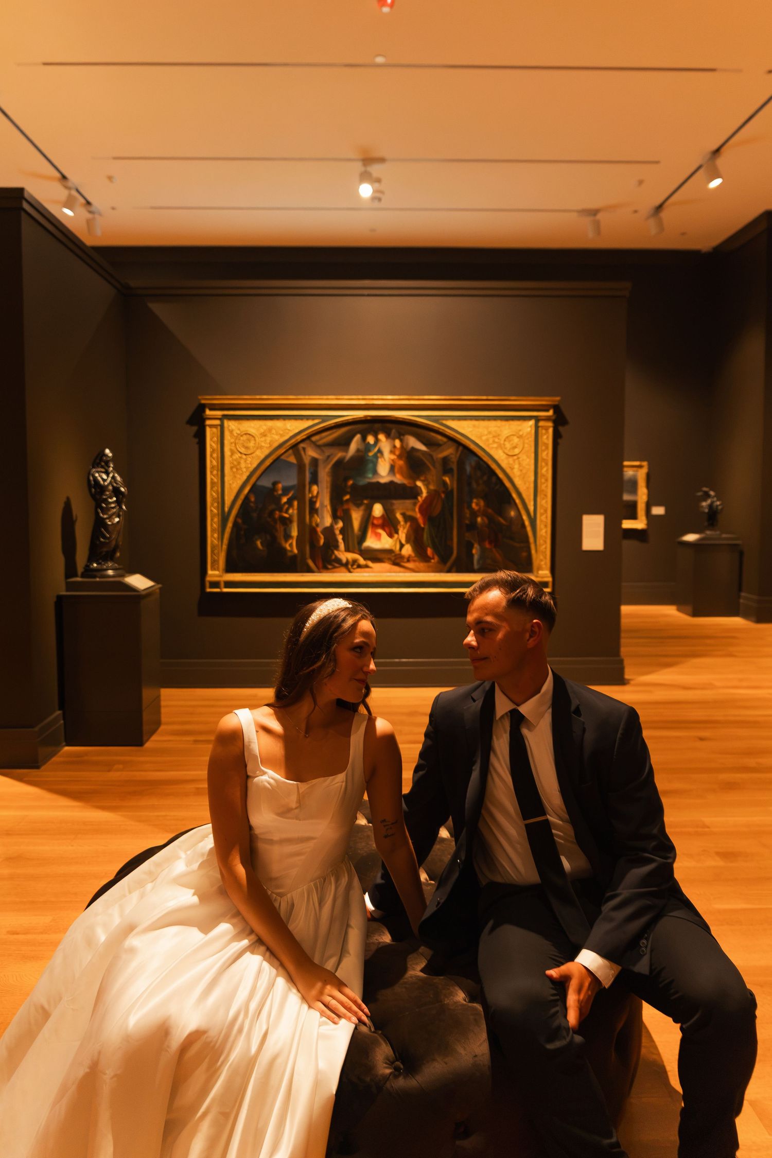 Bride and groom sitting together in an art museum gallery, surrounded by classic paintings and sculptures.