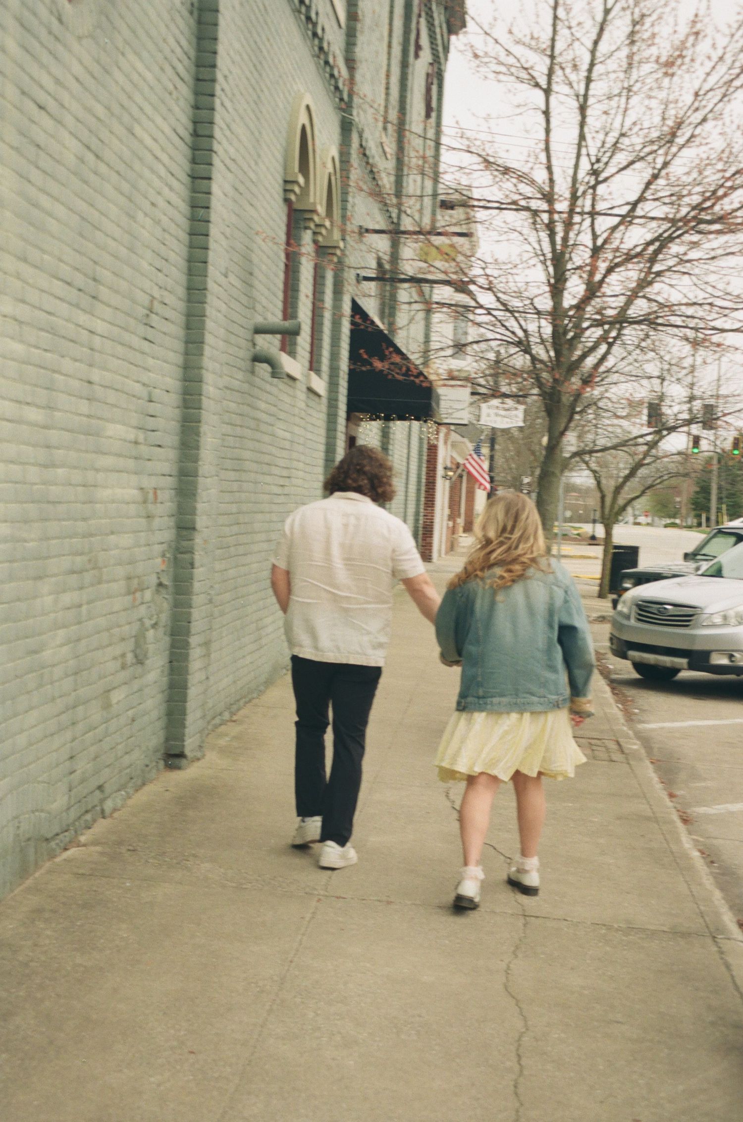 Couple walking hand-in-hand down a city sidewalk, woman in denim jacket and yellow skirt.