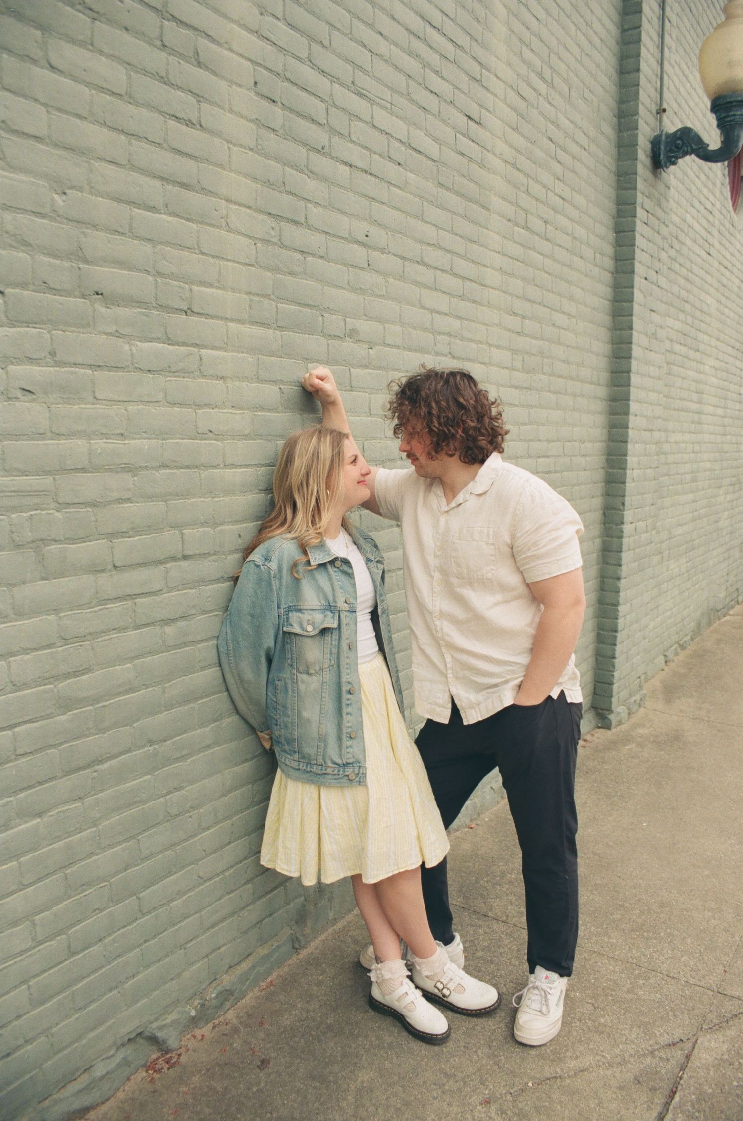 Couple sharing a tender moment against a grey brick wall, woman in yellow skirt and denim jacket.