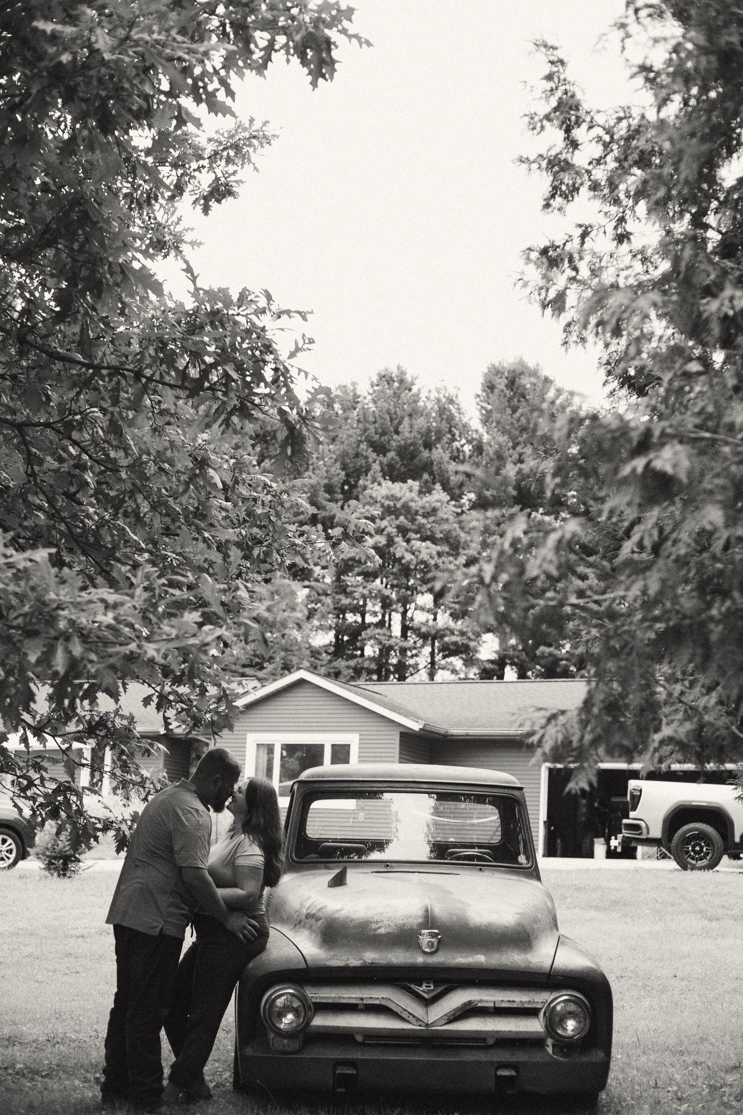Black and white photo of a couple sharing a romantic moment leaning against a vintage Ford pickup truck outdoors.
