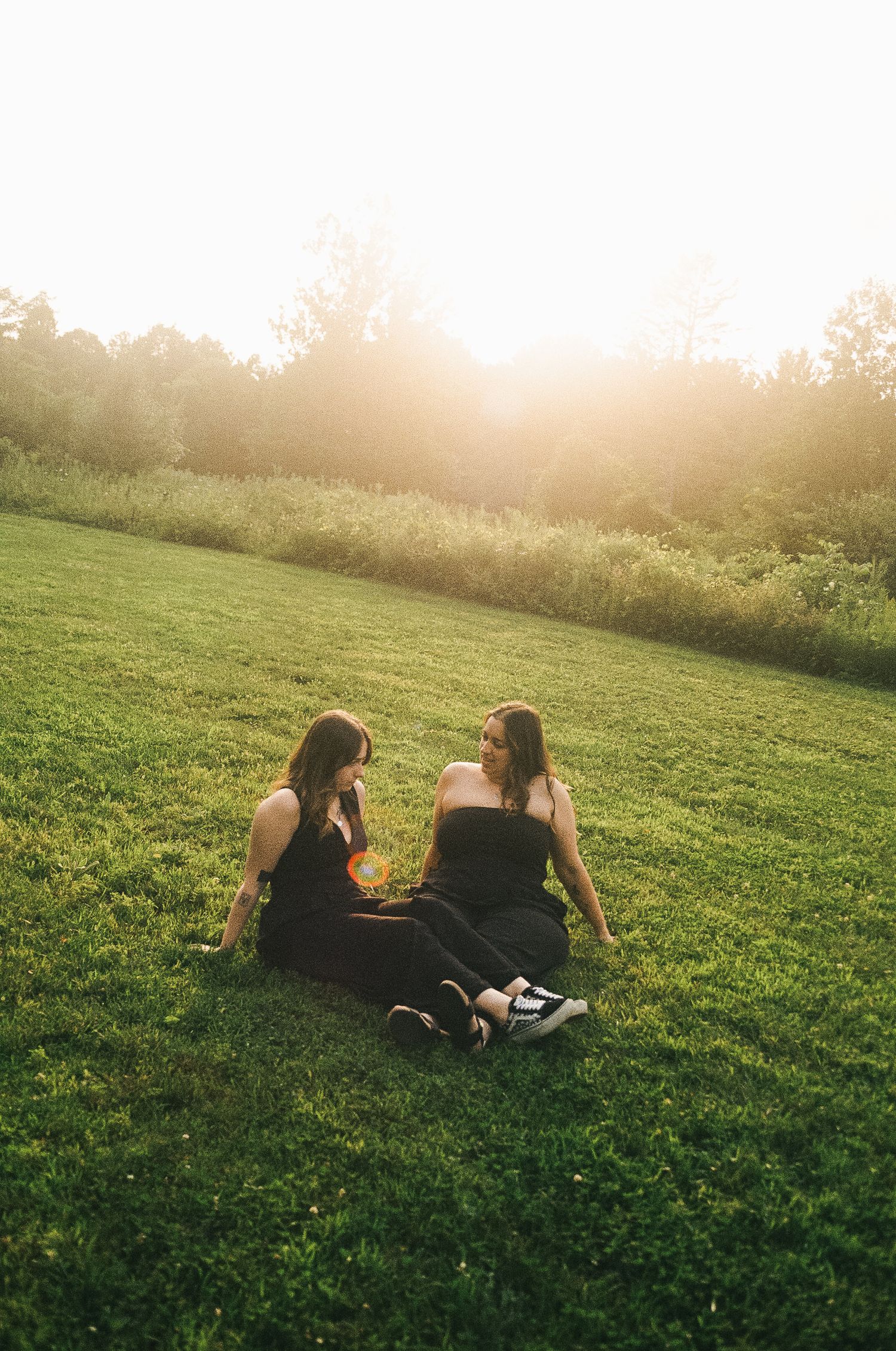 Two young women in black outfits sit on lush green grass chatting during a golden sunset in a scenic outdoor setting.