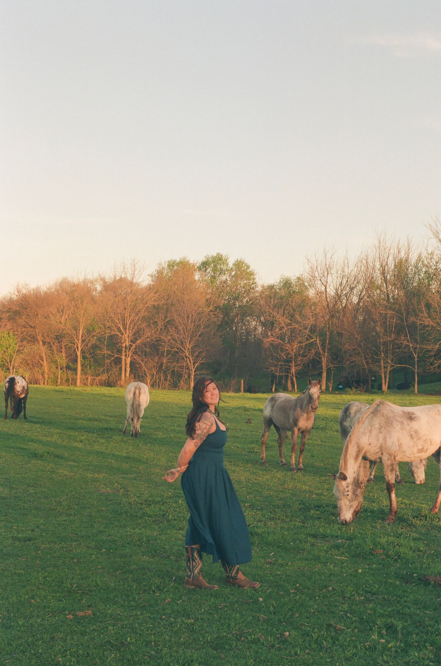 Woman in flowing teal dress stands joyfully among white horses grazing at sunset on green field.