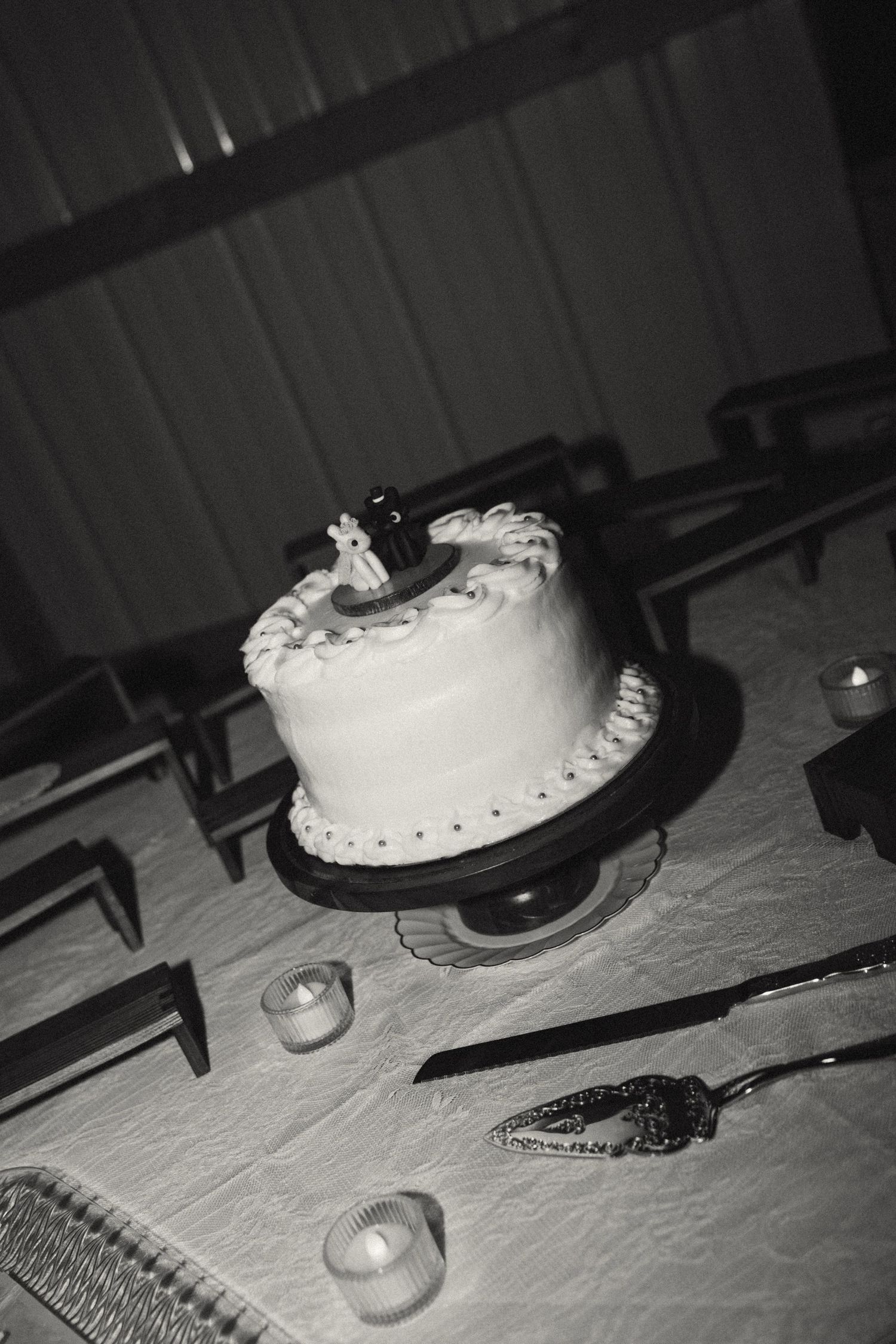 A small white wedding cake with decorative topper sits on a cake stand beside candles in black and white.