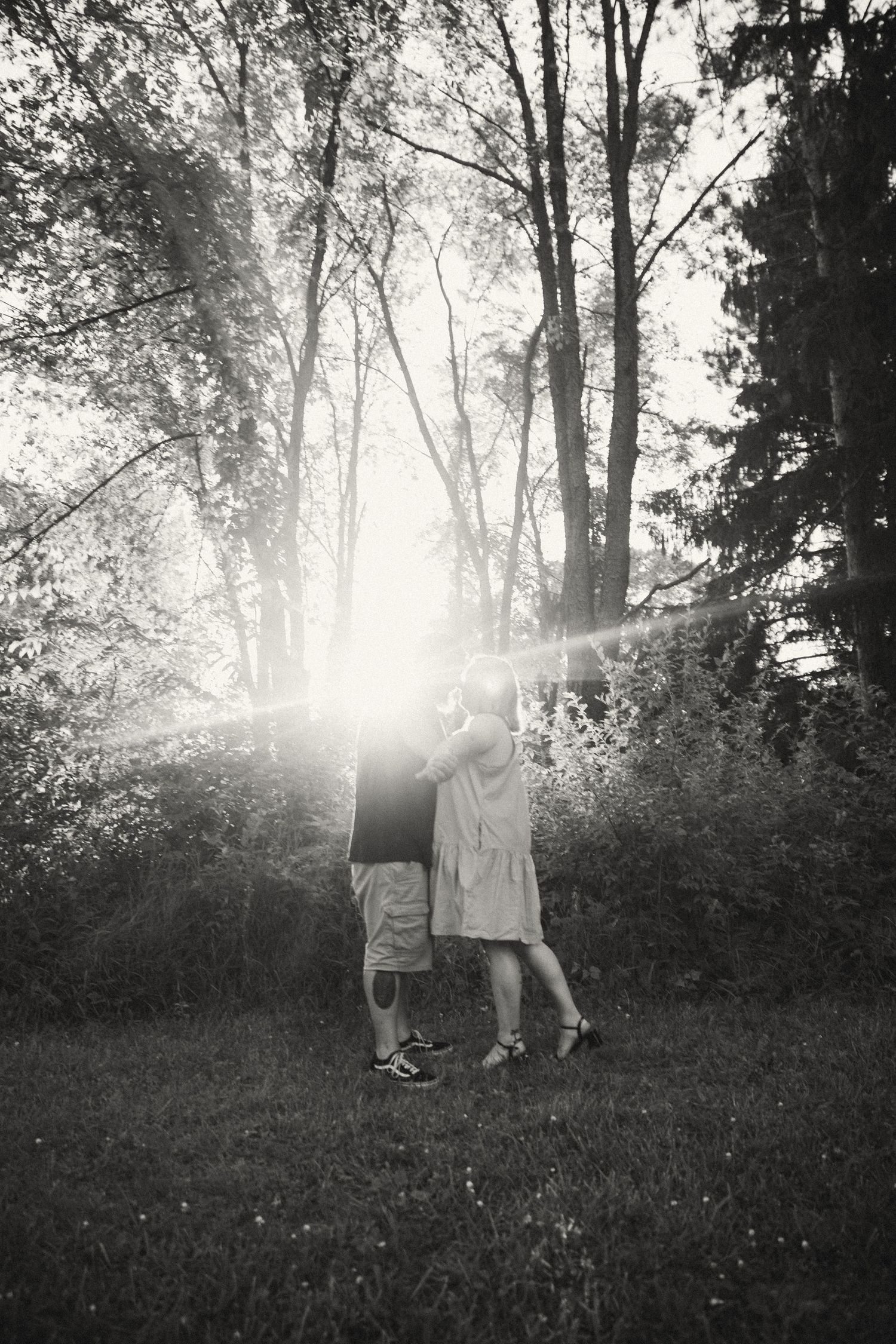 A black and white photo of a couple kissing in a sunlit forest with dramatic lens flare.