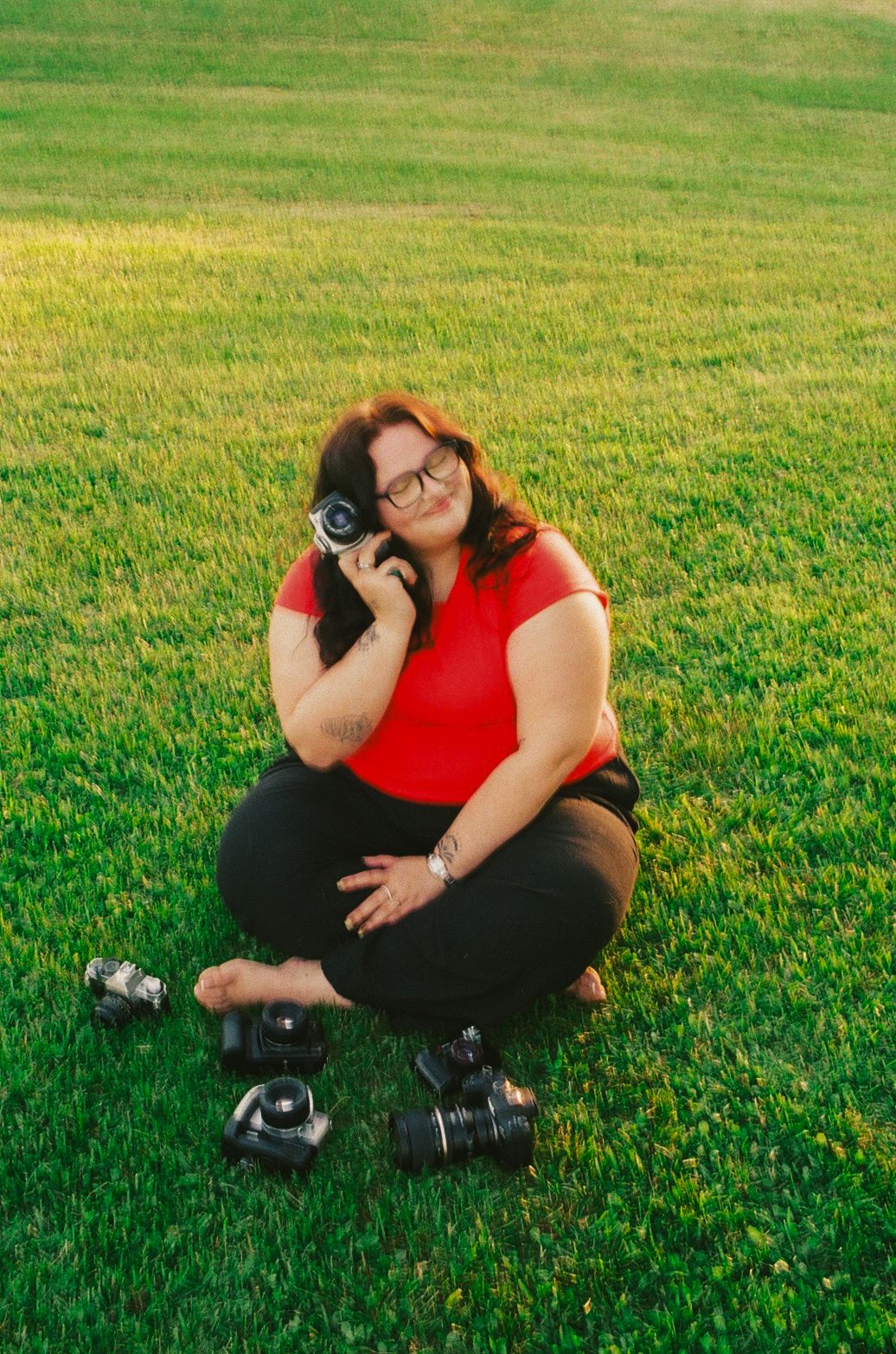 Female photographer crouches on green grass holding a film camera with multiple cameras displayed around her.