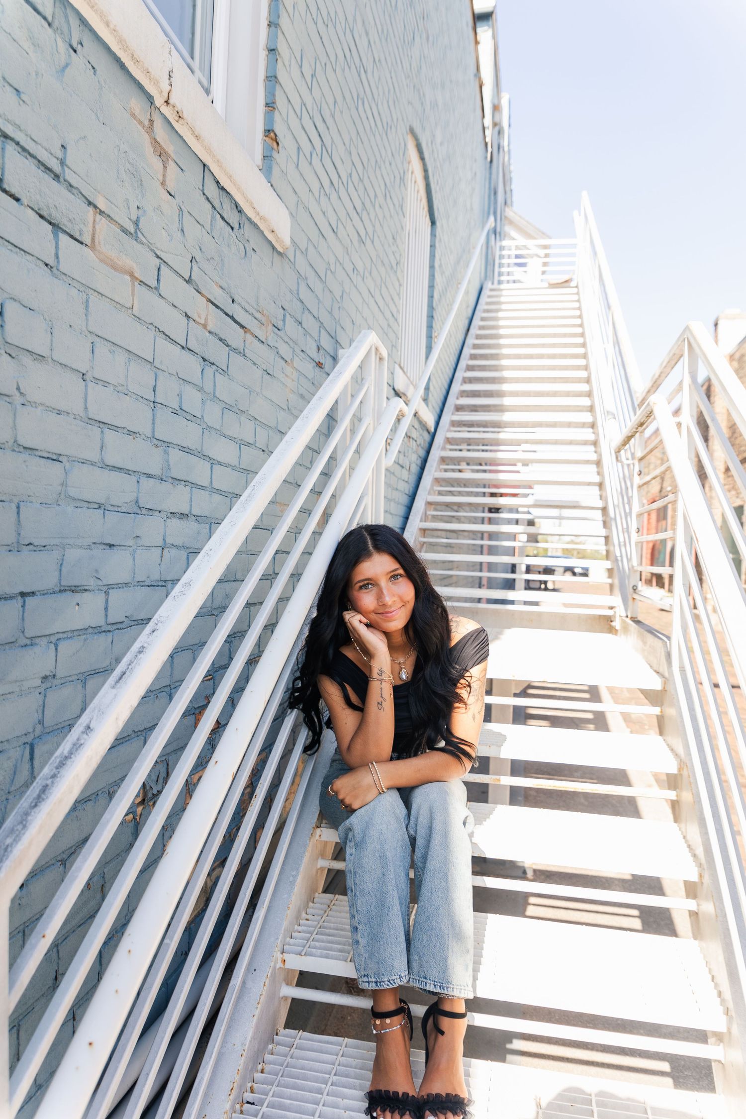 Young woman with long dark hair sitting on white metal outdoor stairs against a brick wall.