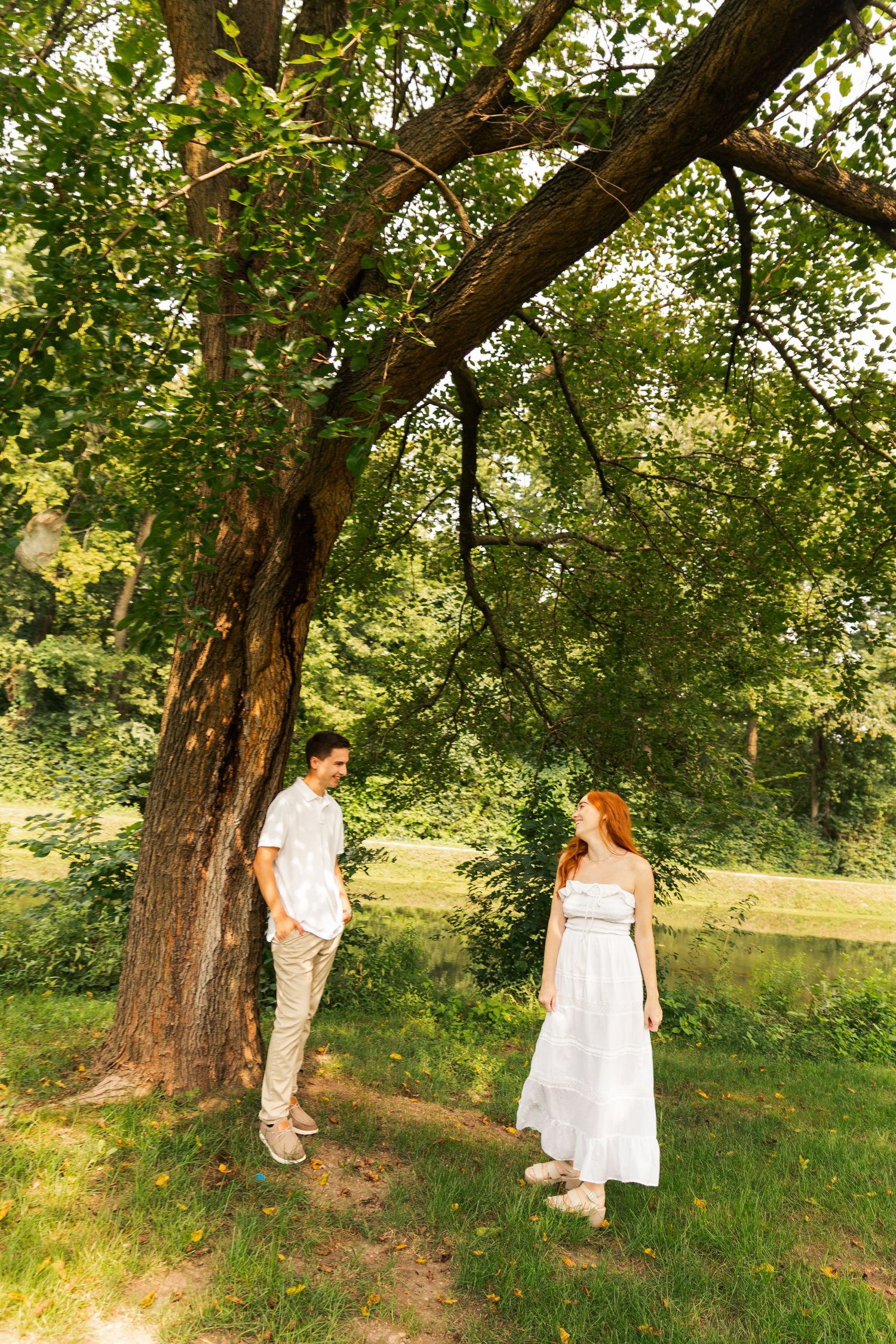 A couple in white outfits stands beneath a large tree in a lush green park on a sunny day.