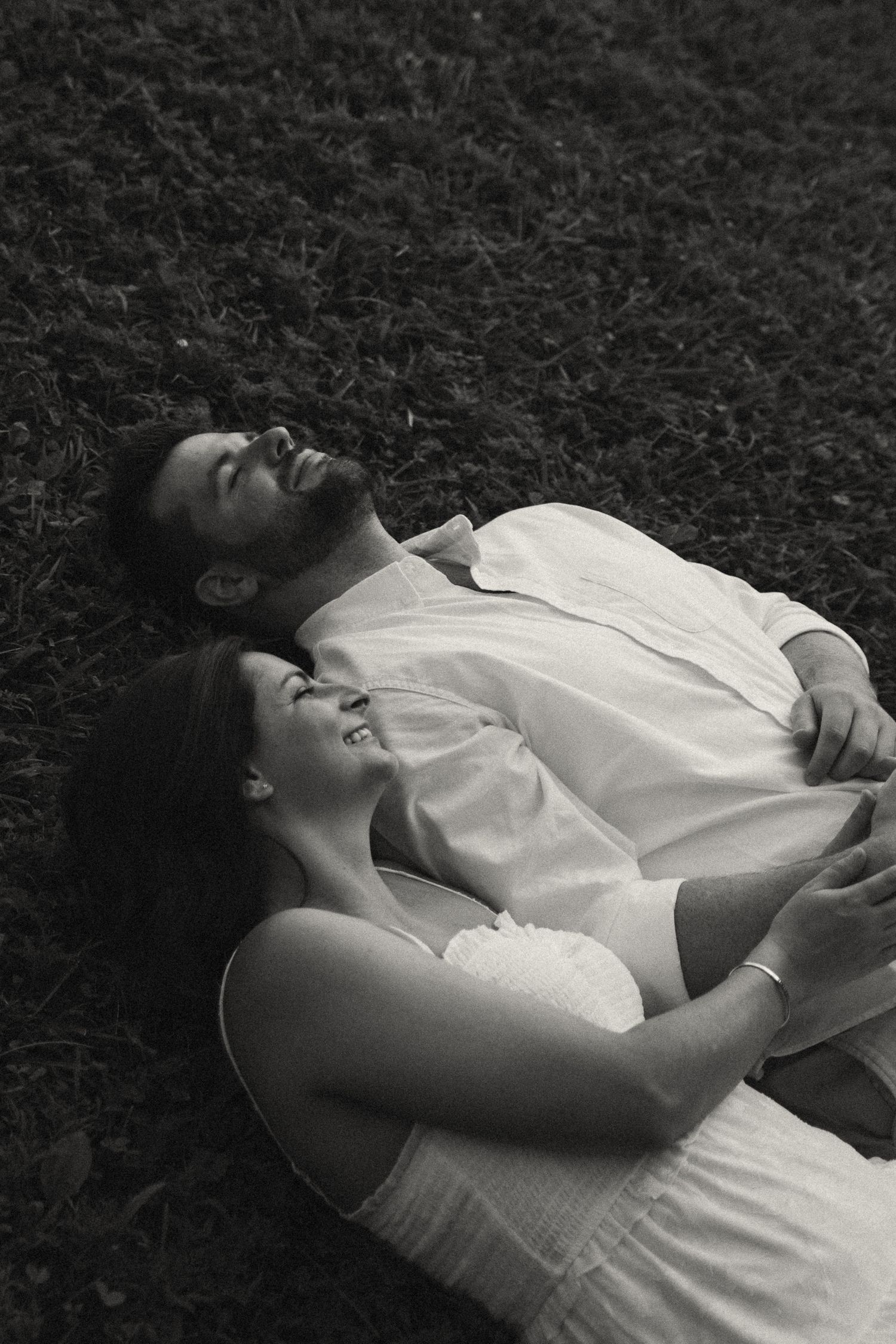 Black and white photo of a smiling couple lying on grass, embracing and looking up joyfully.
