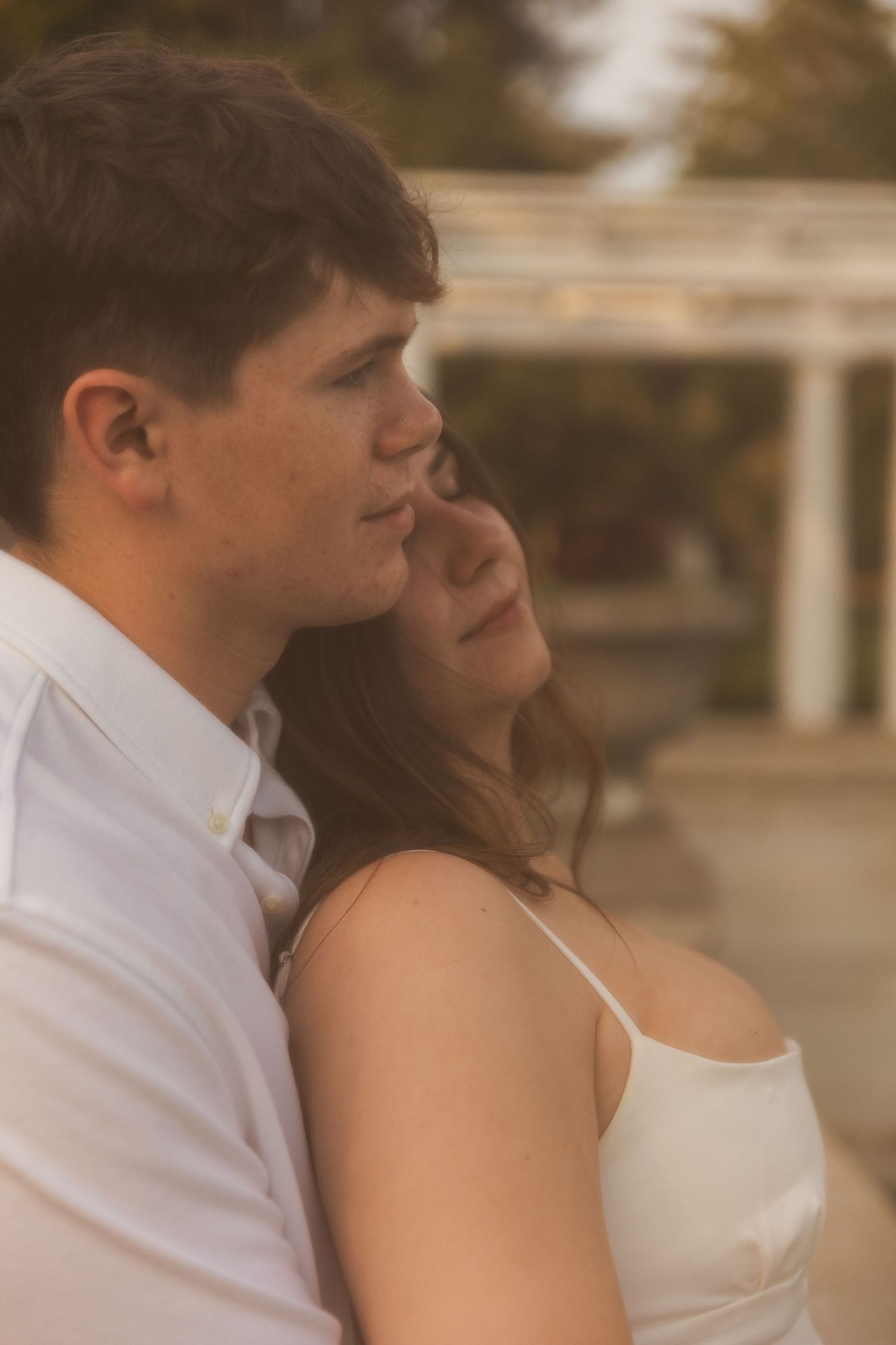 Young couple in white sharing a tender moment outdoors, woman resting her head near mans shoulder at golden hour.
