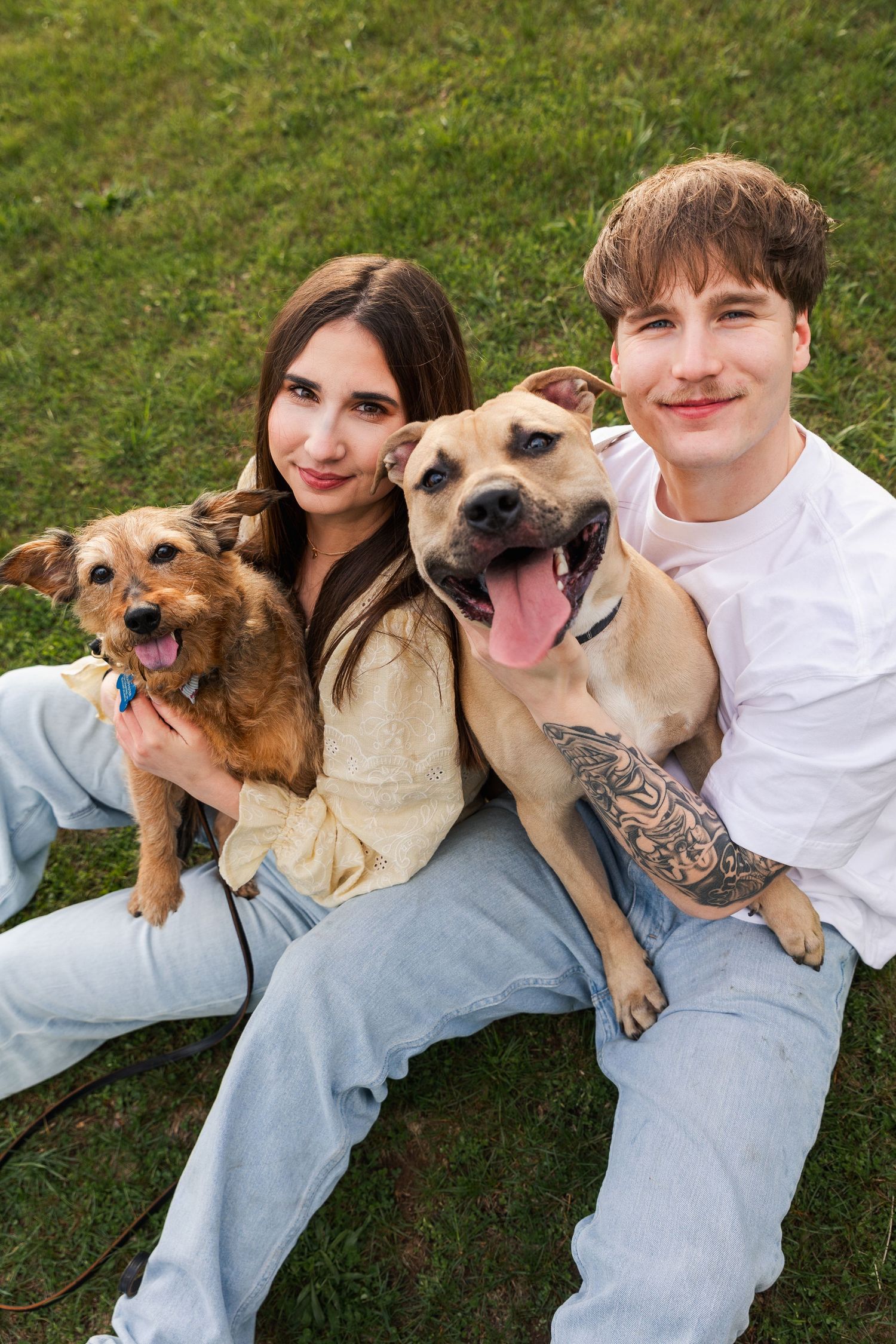 Smiling couple sitting on grass holding two happy dogs, man with tattoos in white shirt.