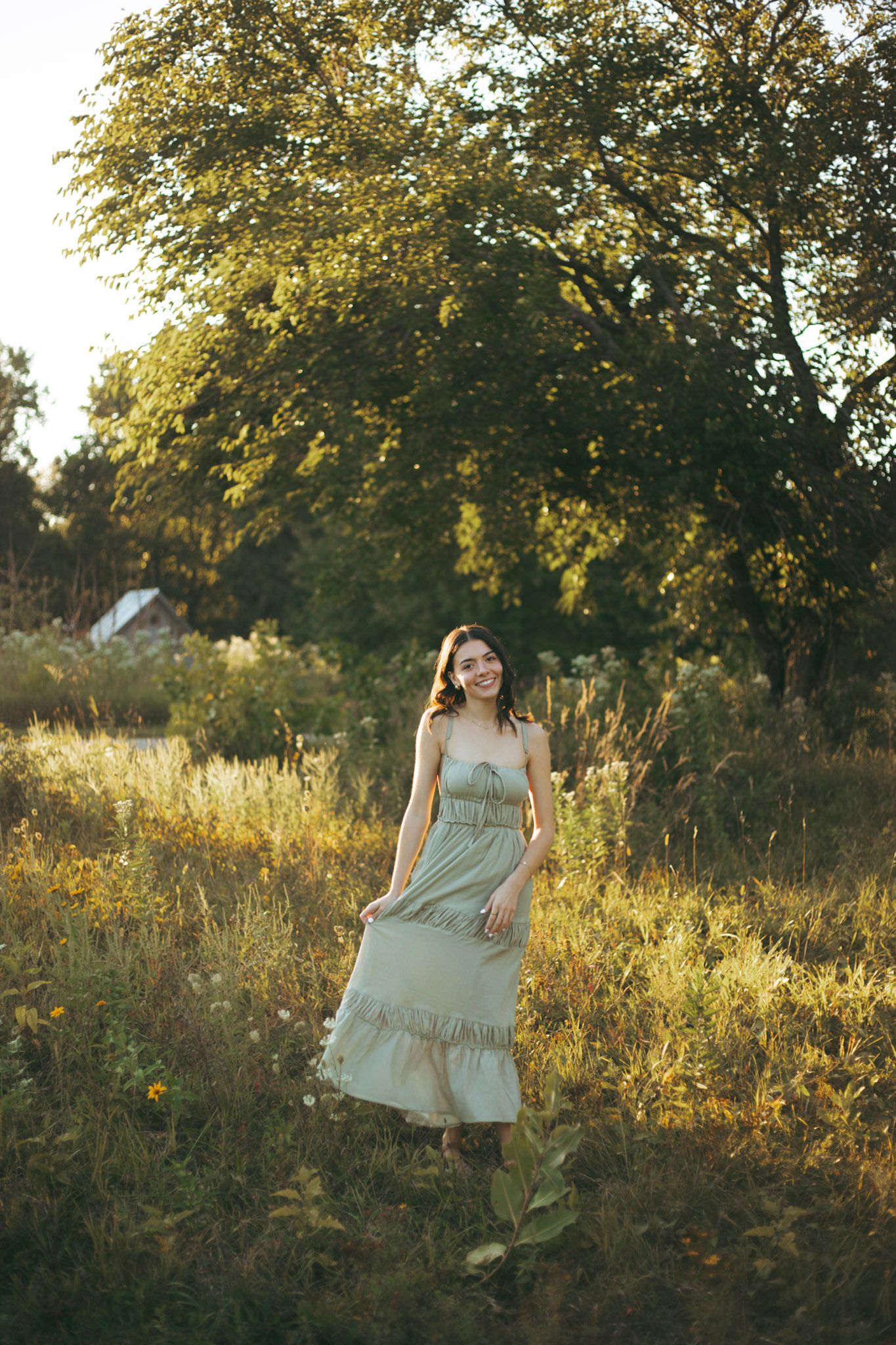 Young woman in sage green sundress walking through golden sunlit meadow with trees in background.