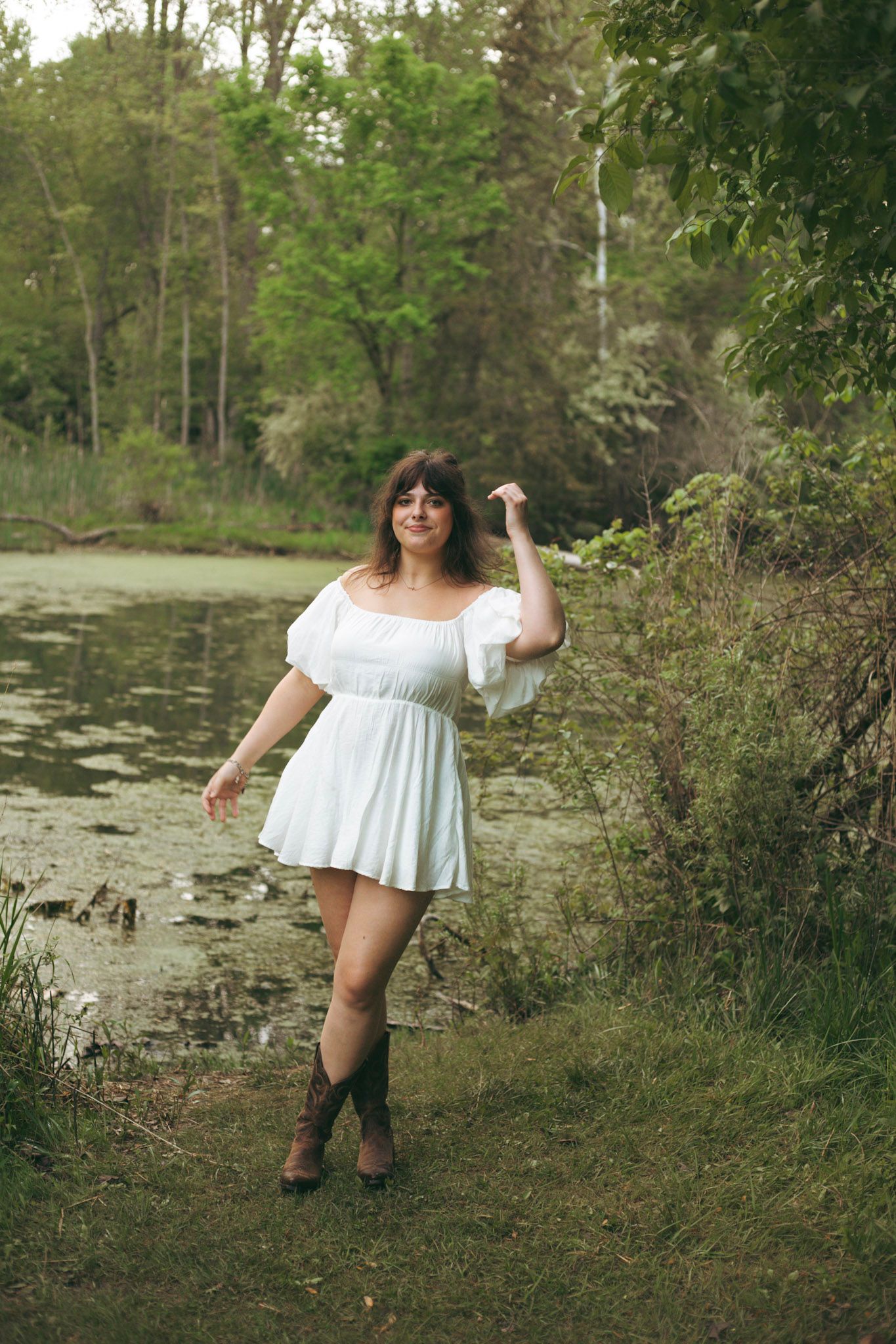 Young woman in white mini dress and cowboy boots posing beside a tranquil pond in a forest.