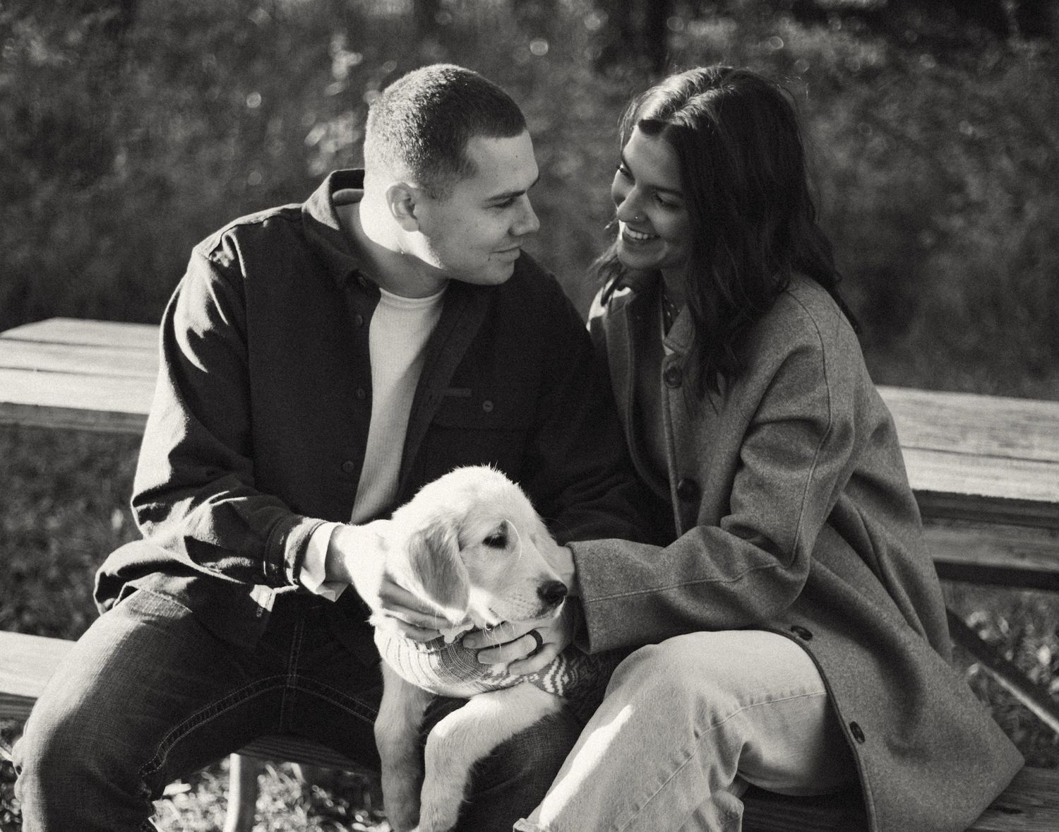 A black and white photo of a couple sitting on a bench, smiling at each other while petting a white puppy.