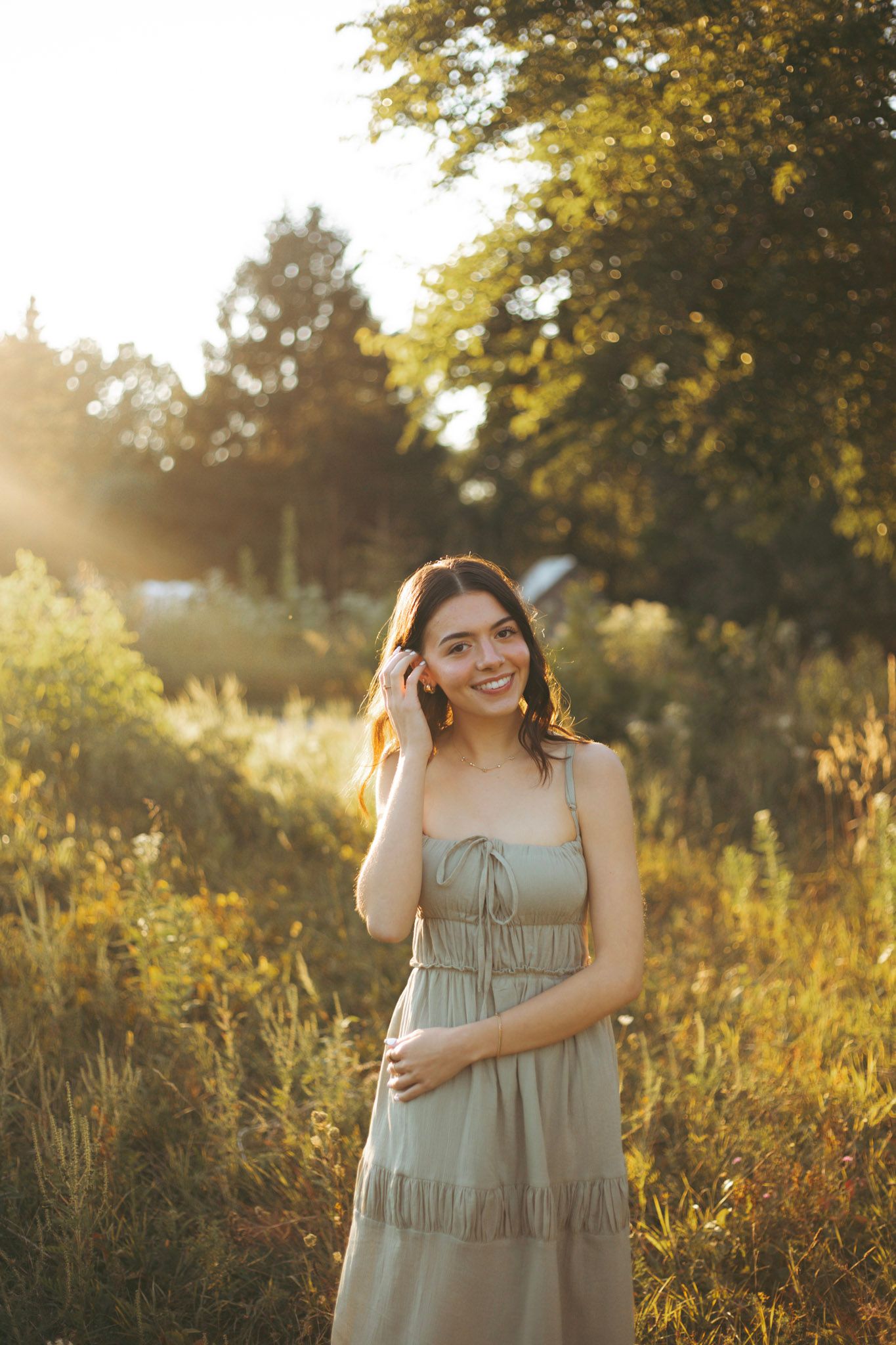 Smiling young woman in sage green sundress standing in sunlit field with warm golden hour lighting.