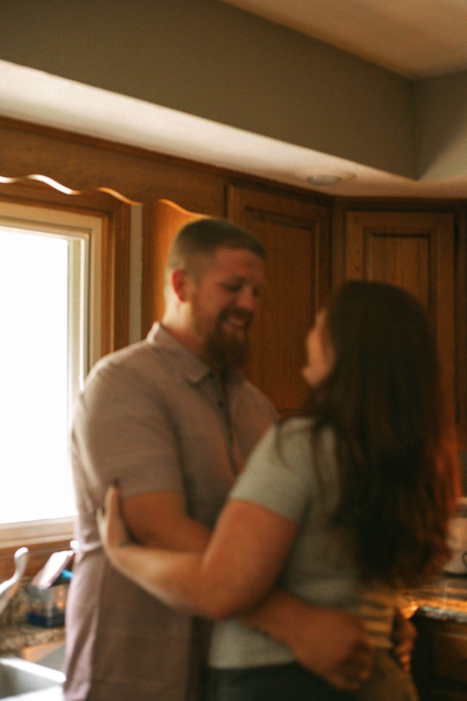 A bearded man and woman share a warm embrace while dancing in a sunlit kitchen with wooden cabinets.