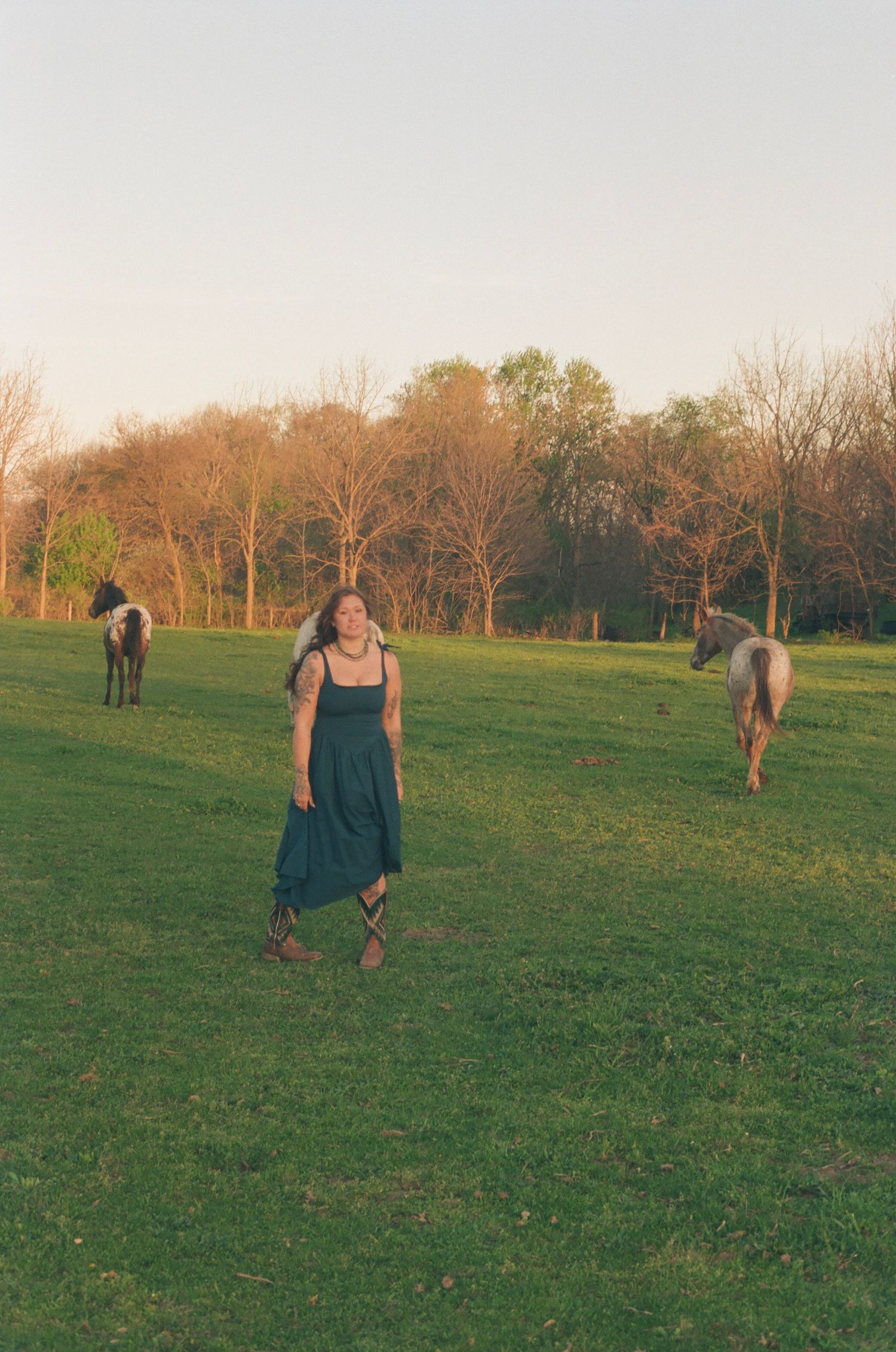 Woman in teal dress walks freely through green pasture with horses grazing at golden hour.