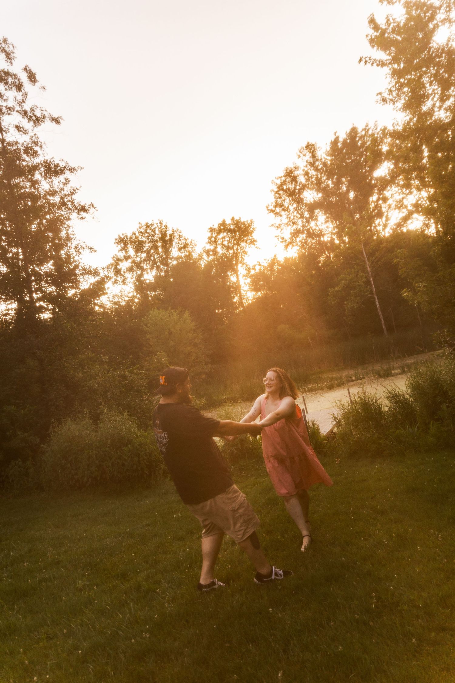Couple dancing joyfully on green grass at golden sunset with trees and pond in background.