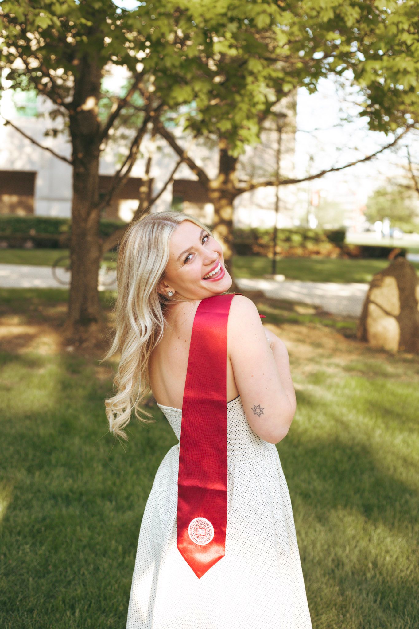Smiling blonde graduate in white dress with red graduation sash posing outdoors on a sunny day.