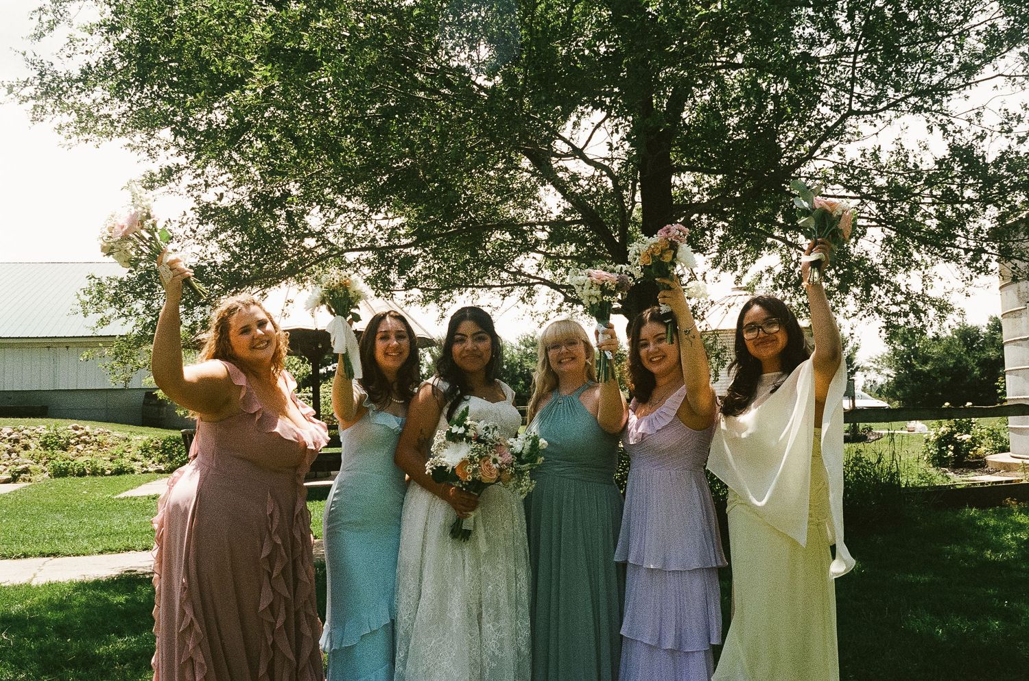 Bride with five bridesmaids in colorful mismatched dresses celebrating outdoors raising bouquets under sunny sky.