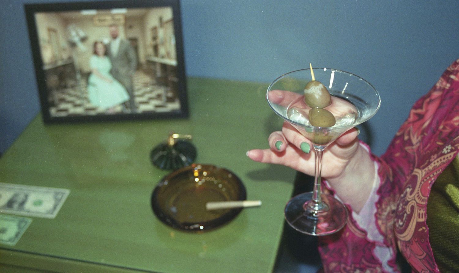 Hand holding martini glass near vintage wedding photo frame on green table with ashtray.