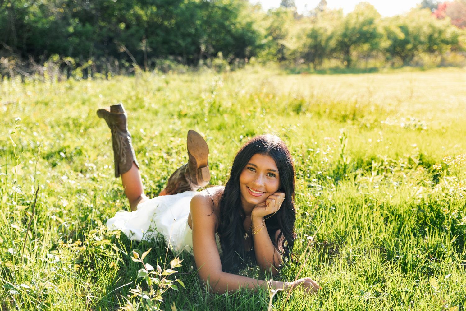 Smiling brunette woman lying in a sunny grass field wearing a white dress and boots.
