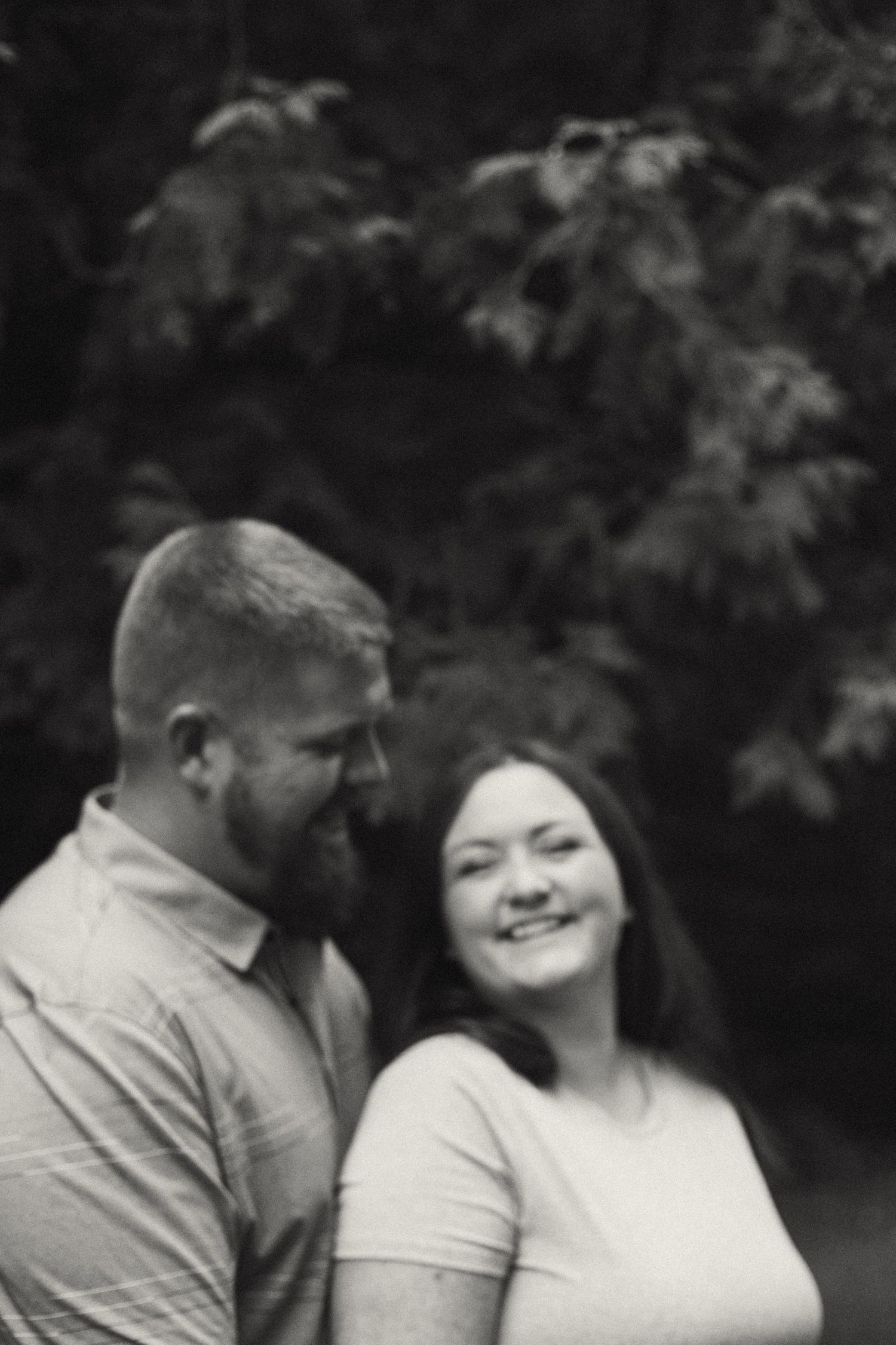 Black and white portrait of a smiling couple standing close together outdoors beneath lush green trees.