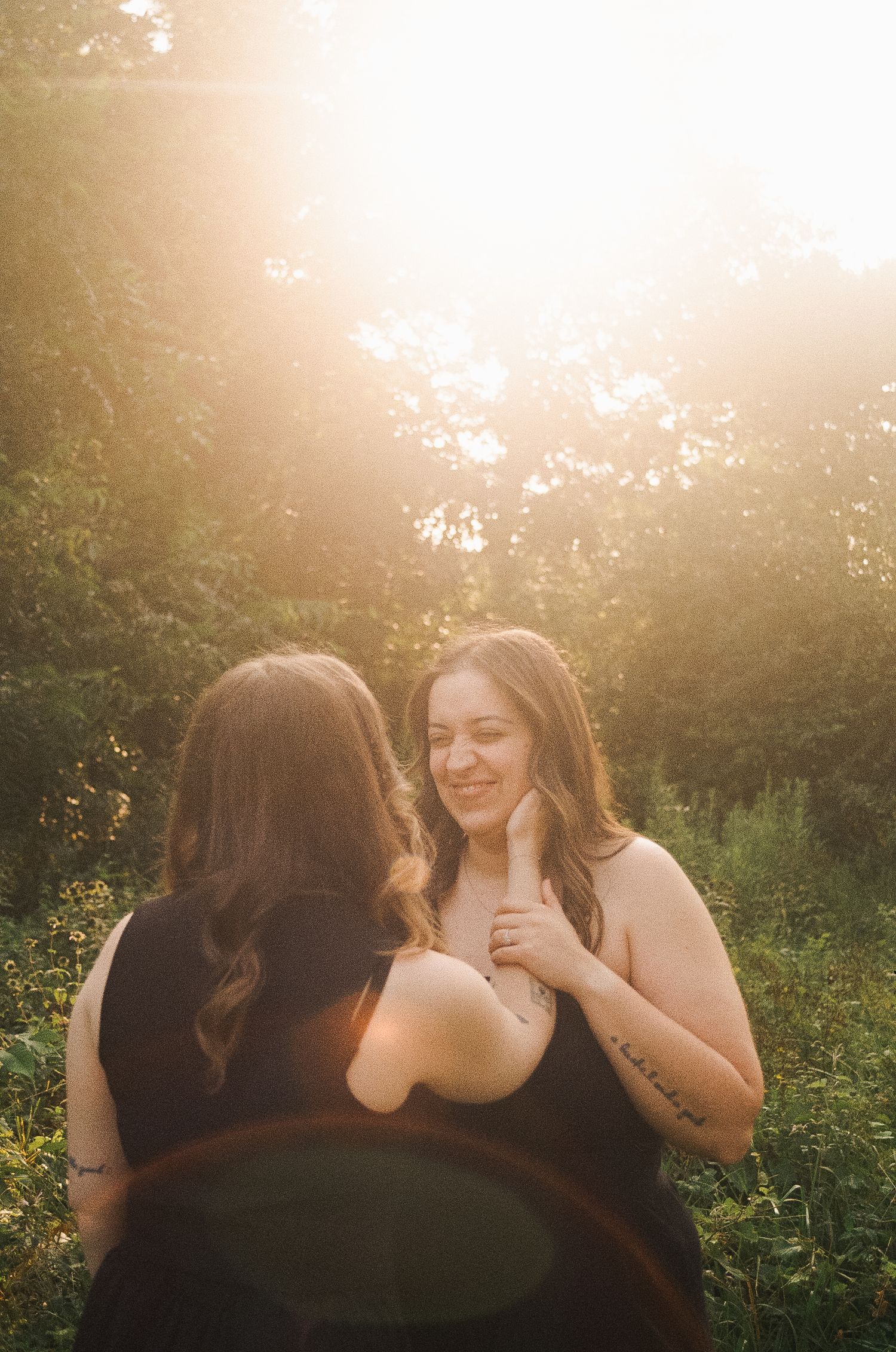 Two women share a joyful moment outdoors, bathed in golden sunlight with lush green trees in the background.