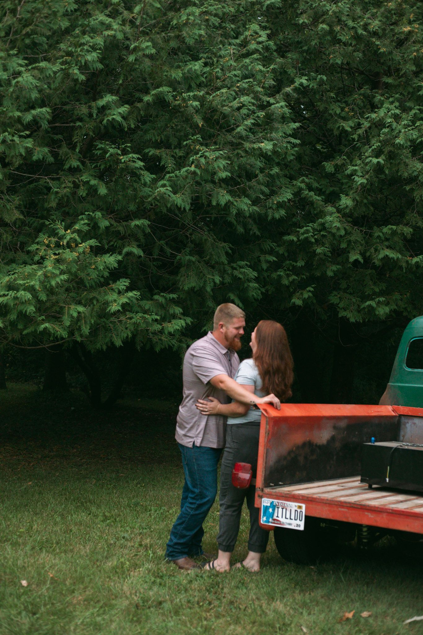Couple sharing a romantic embrace near a vintage truck surrounded by lush green trees.