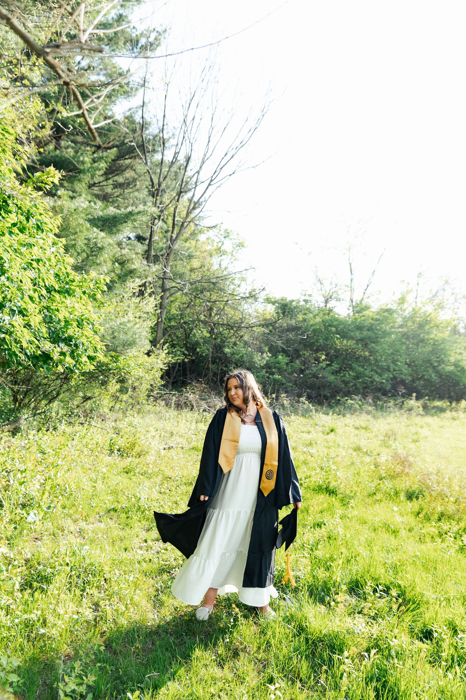 Woman in graduation gown and gold stole standing in a sunny green meadow.
