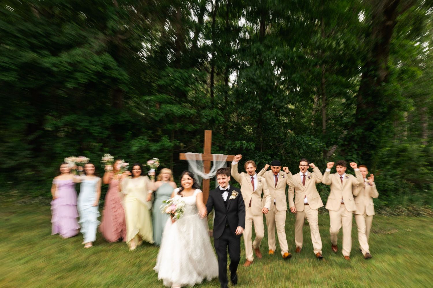 Wedding party celebrating outdoors near a wooden cross surrounded by lush green trees.