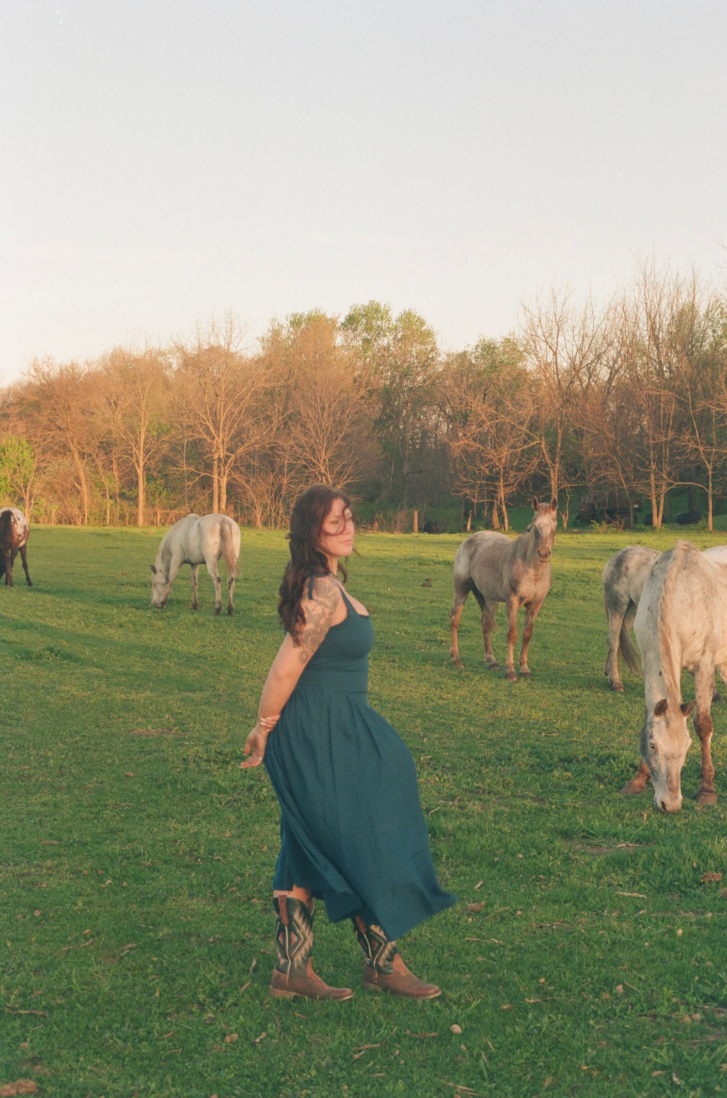 Smiling woman in teal dress poses with grazing white horses in a warm sunset pasture.