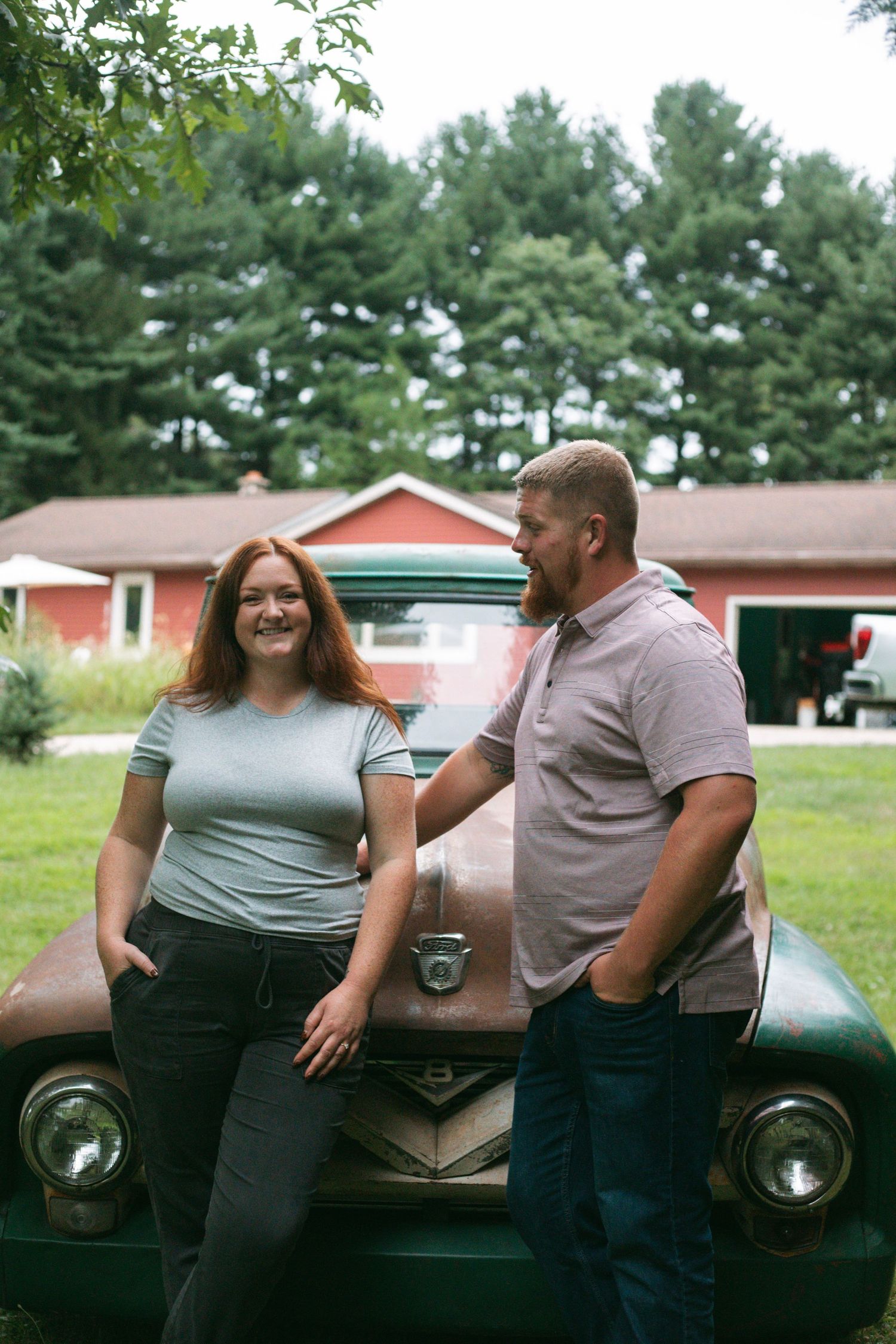 A smiling couple leans against a rustic green vintage truck in front of a red barn surrounded by trees.