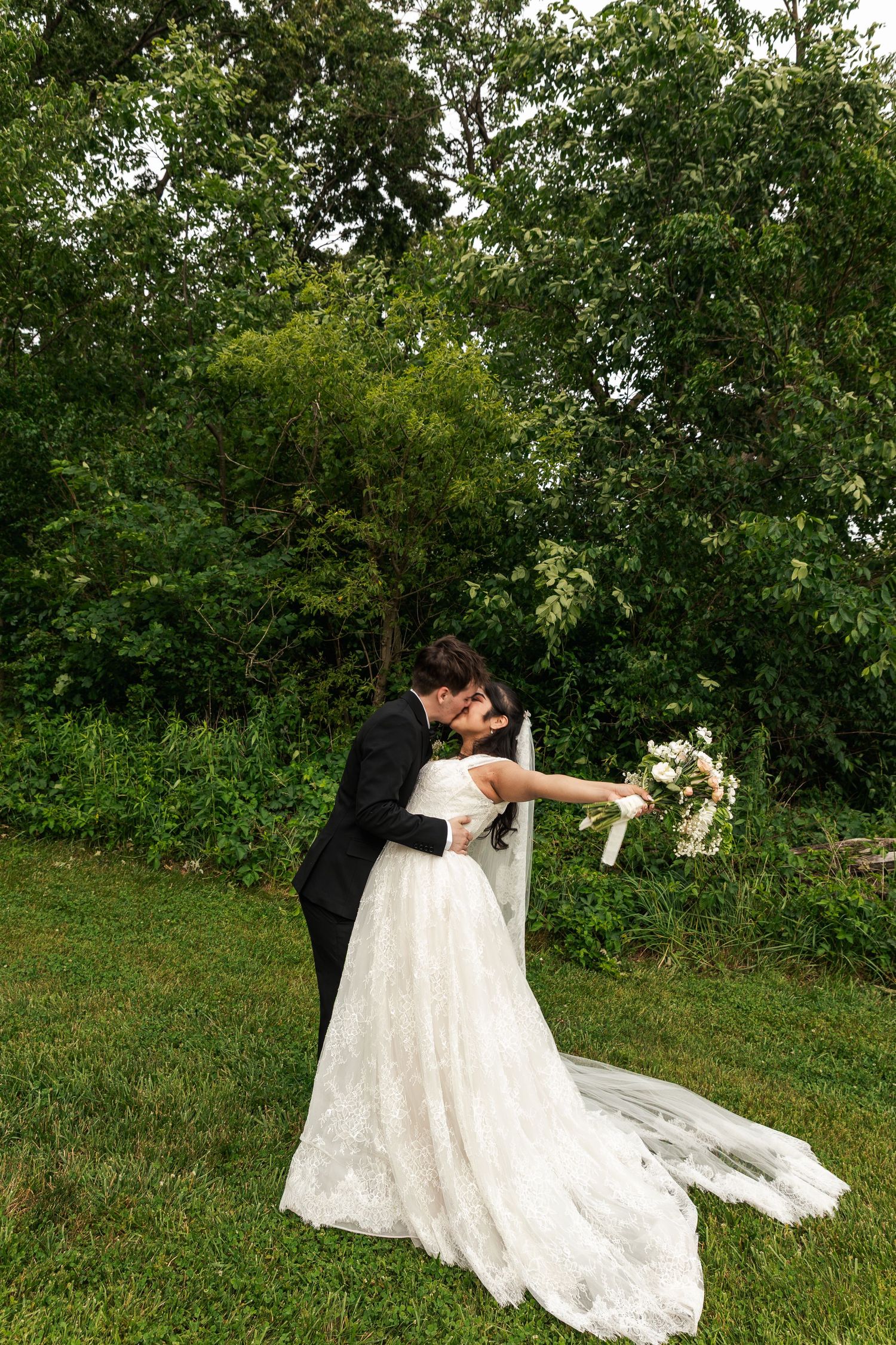 A bride and groom share a romantic dip kiss outdoors on green grass, surrounded by lush trees, as she holds a white bouquet.
