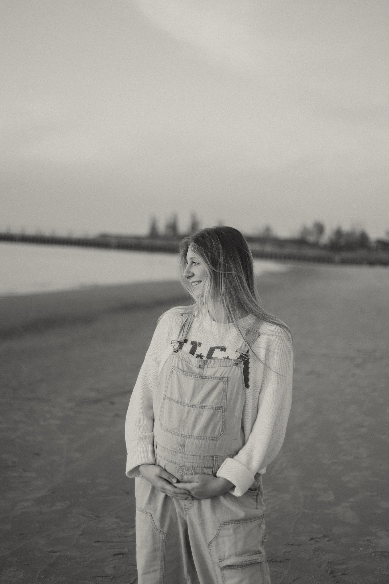 Black and white portrait of a pregnant woman in denim overalls smiling on a sandy beach.