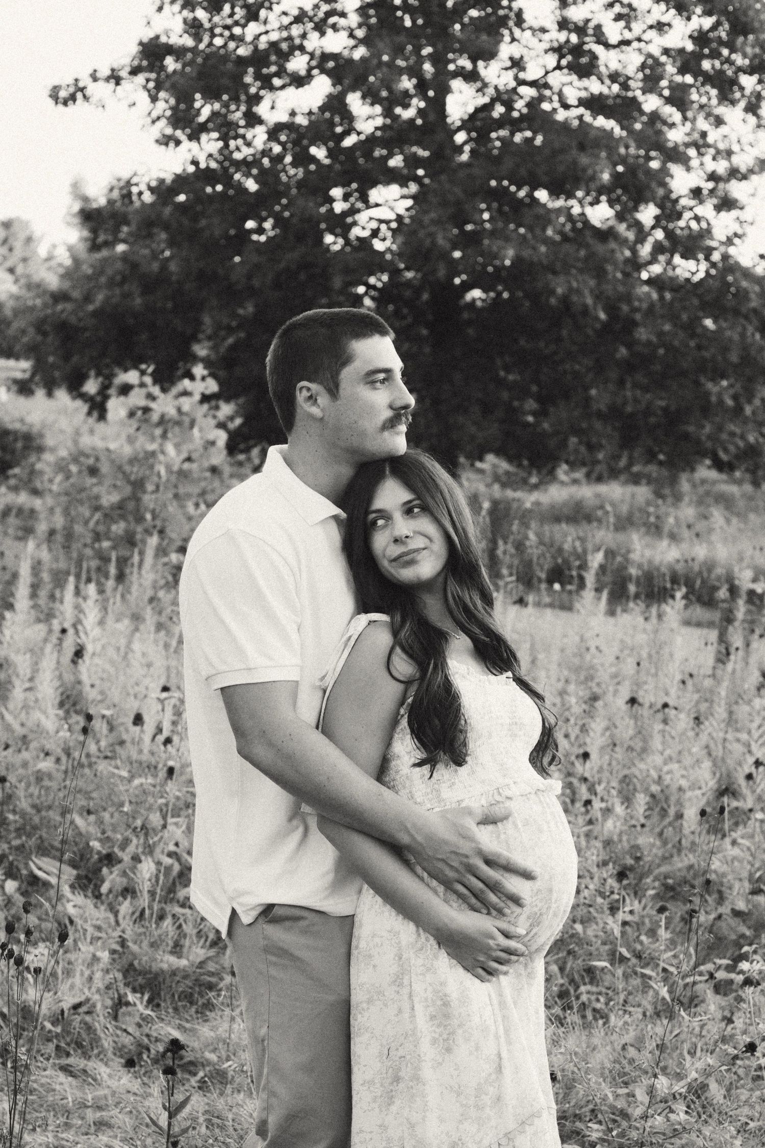 Black and white maternity portrait of couple embracing in wildflower field, man cradling pregnant belly.