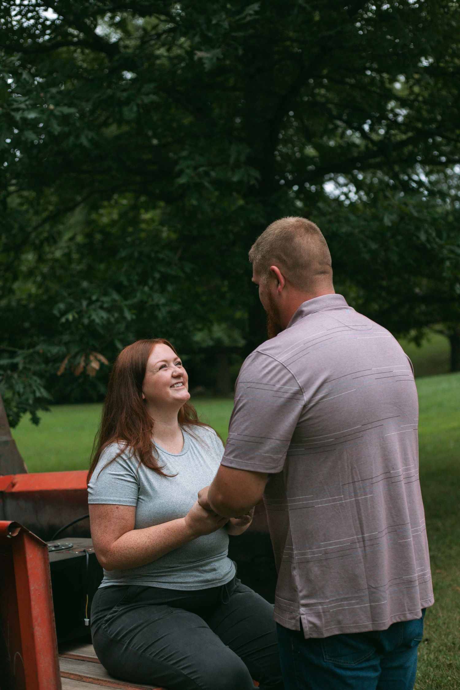 A laughing redheaded woman sits on a red truck tailgate holding hands with a man in a lush green park.