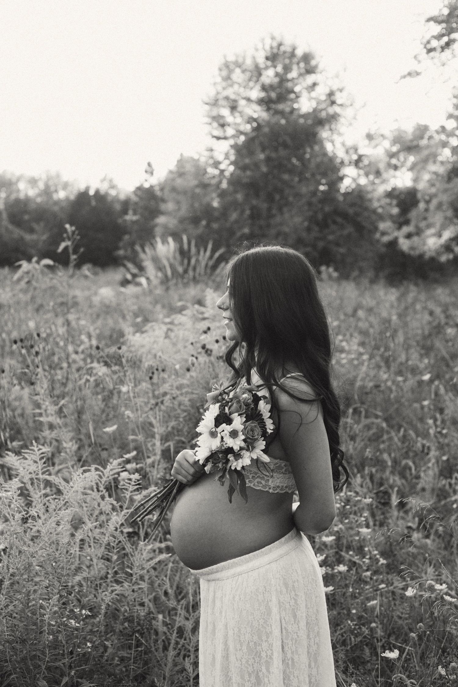 Black and white rear view of pregnant woman holding wildflower bouquet in a meadow.