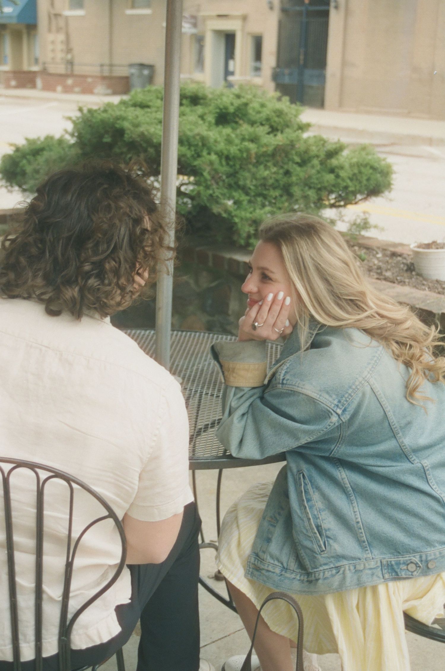 Couple at outdoor cafe table, woman in denim jacket leaning in closely while showing engagement ring.