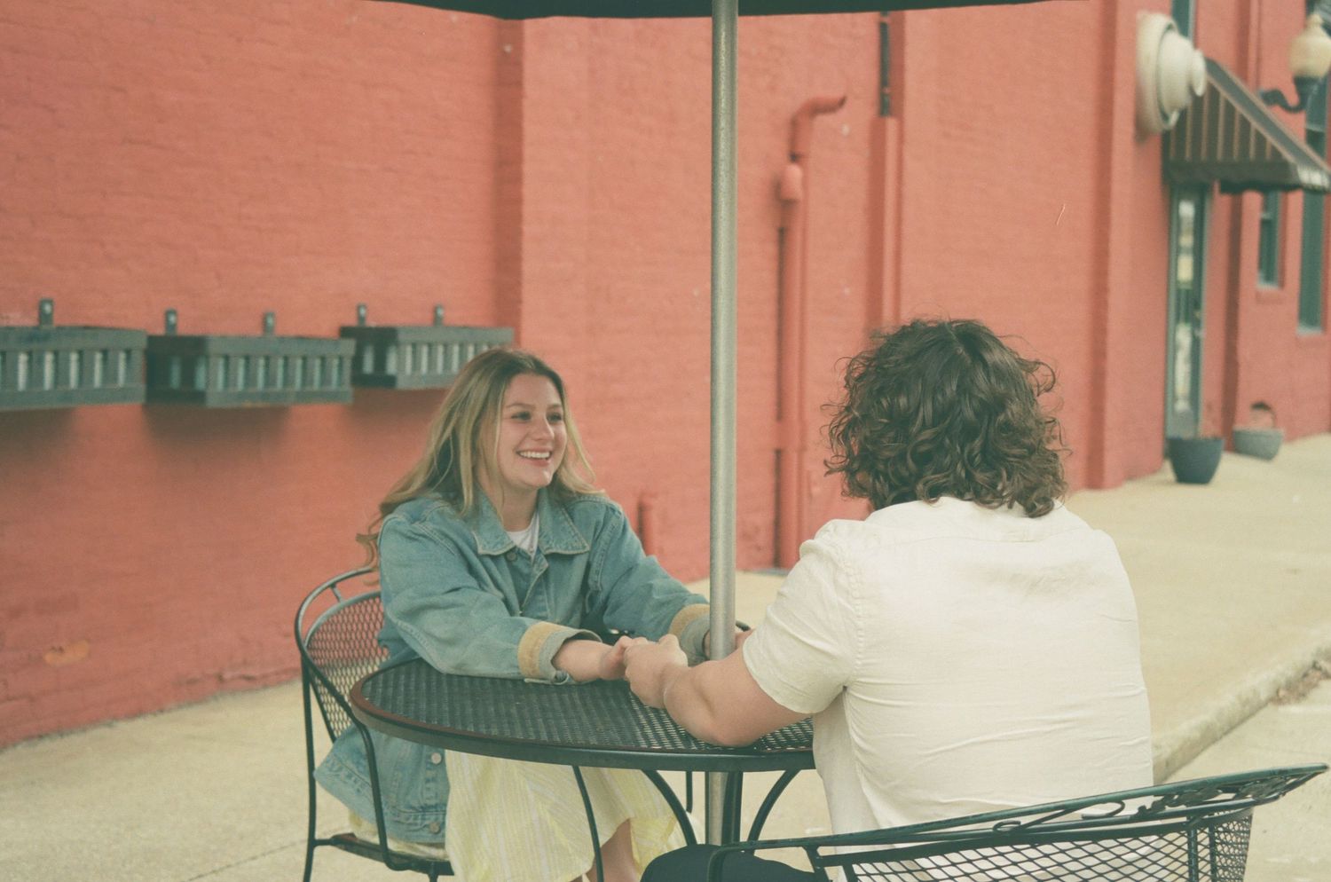 A smiling woman in a denim jacket sits across from a man at an outdoor patio table holding hands.