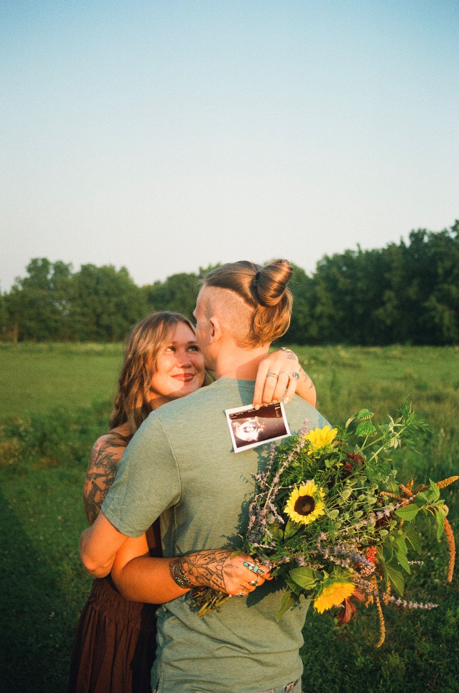 Couple embracing in a field holding a bouquet of sunflowers and wildflowers with a sonogram photo tucked inside.